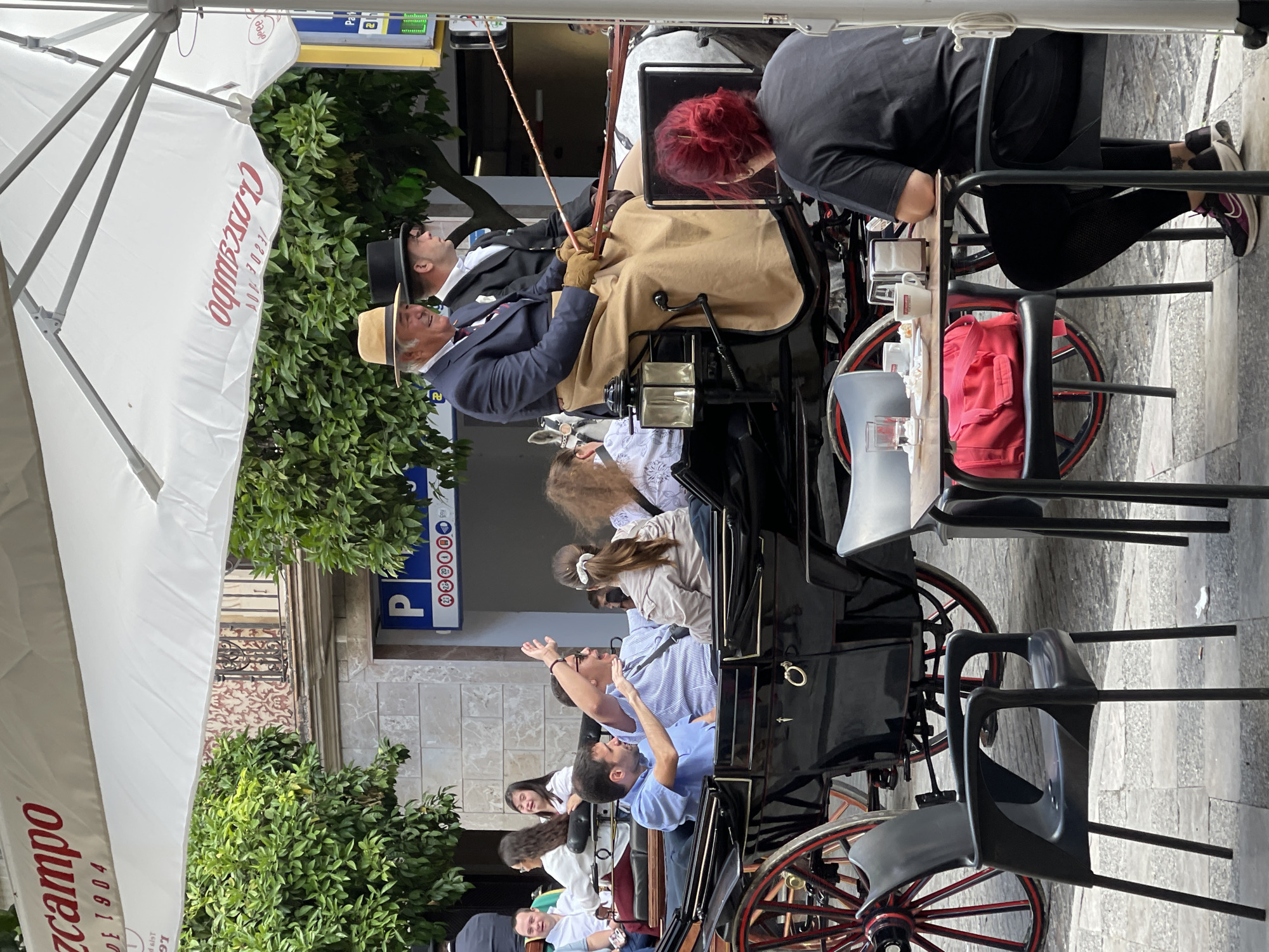 The image shows a horse-drawn carriage in Jerez De La Frontera, Spain. The carriage is occupied by several people, including the driver who is wearing a suit, gloves, and a straw hat and an assistant with red hair. Other passengers are also visible in the carriage's seating area. The carriage is on a street with tables and chairs from a cafe or restaurant. The street is paved and lined with buildings and trees. There is a large umbrella with a company name, "Cruzcampo", written on it. A sign with a blue "P" is also visible. The scene seems to depict a tourist activity, such as a carriage tour.