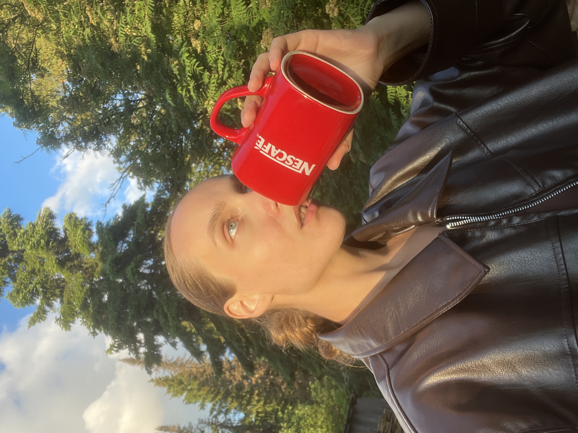 This portrait-oriented image, taken in Sharkan, Russia, features a person holding a red Nescafé mug outdoors. The subject, a young adult with light hair pulled back and light-colored eyes, is seen from a side profile, looking upwards and to the right. She is wearing a dark, possibly leather-look, jacket with visible stitching and a zipper detail on the lapel.
The activity shows the person holding the bright red mug with her right hand close to her mouth, appearing to be taking a sip from the dark liquid inside. The word "NESCAFE" is clearly visible in white capital letters on the side of the mug.
The setting is an outdoor, natural environment. The background is dominated by lush green trees, including what appears to be conifers and other leafy trees. Patches of clear blue sky with fluffy white clouds are visible between the tree canopy, indicating fair weather. The scene is illuminated by warm, golden light, highlighting the subject's face and jacket, suggesting it is either late afternoon or early morning (golden hour).