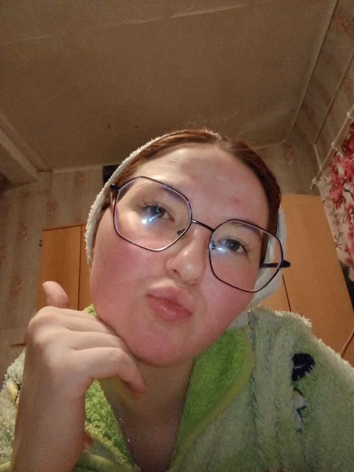 A close-up, upward-angle indoor photograph depicts a female subject from the upper torso, framed against an interior background. The subject has reddish-brown hair, partially covered by a white, towel-like headband, and wears oversized, dark-rimmed, geometric-shaped prescription glasses. Her face exhibits a generalized reddening, particularly on the cheeks and forehead, and her lips are pursed forward. She is attired in a textured, light green garment, consistent with a robe or a fuzzy top, which features darker green accents or patterns. Her right hand (viewer's left) is positioned under her chin, with the thumb extended vertically in a gesture. Bright light reflections are prominently visible on the lenses of her glasses. The background consists of a light-colored ceiling. To the left of the subject, a wall covered with patterned wallpaper is visible. To the right, a dark, vertical piece of furniture or wall section is partially obscured by a curtain or fabric displaying a red and white floral pattern. The precise location in the city of Старый Шагирт, Russia, cannot be determined from the visual evidence presented in the image.