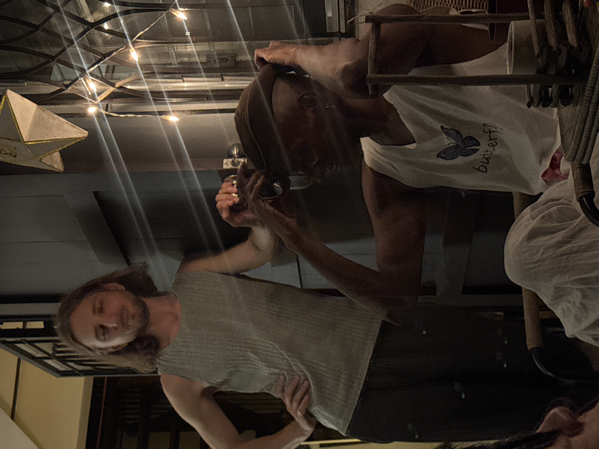 Two people are the subjects in this nighttime photo, likely taken in Chiang Mai, Thailand. The foreground features a person with long, light brown hair and a beard, wearing a light grey, textured sleeveless shirt. They appear to be looking down and slightly to their right. Behind them, a second person with dark skin is partially visible, wearing a white tank top with a blue butterfly design and the handwritten text "butterf!". Their hands are near their head, suggesting they are adjusting their hair or clothing.
The setting appears to be indoors or a covered outdoor space with a reflective ceiling and decorative string lights creating a festive atmosphere. A star-shaped lantern hangs from the ceiling on the left side of the frame. The overall lighting is dim, with the string lights providing points of illumination. The image is oriented sideways, making the subjects appear to be standing or sitting in a vertical orientation. The mood is casual and relaxed.