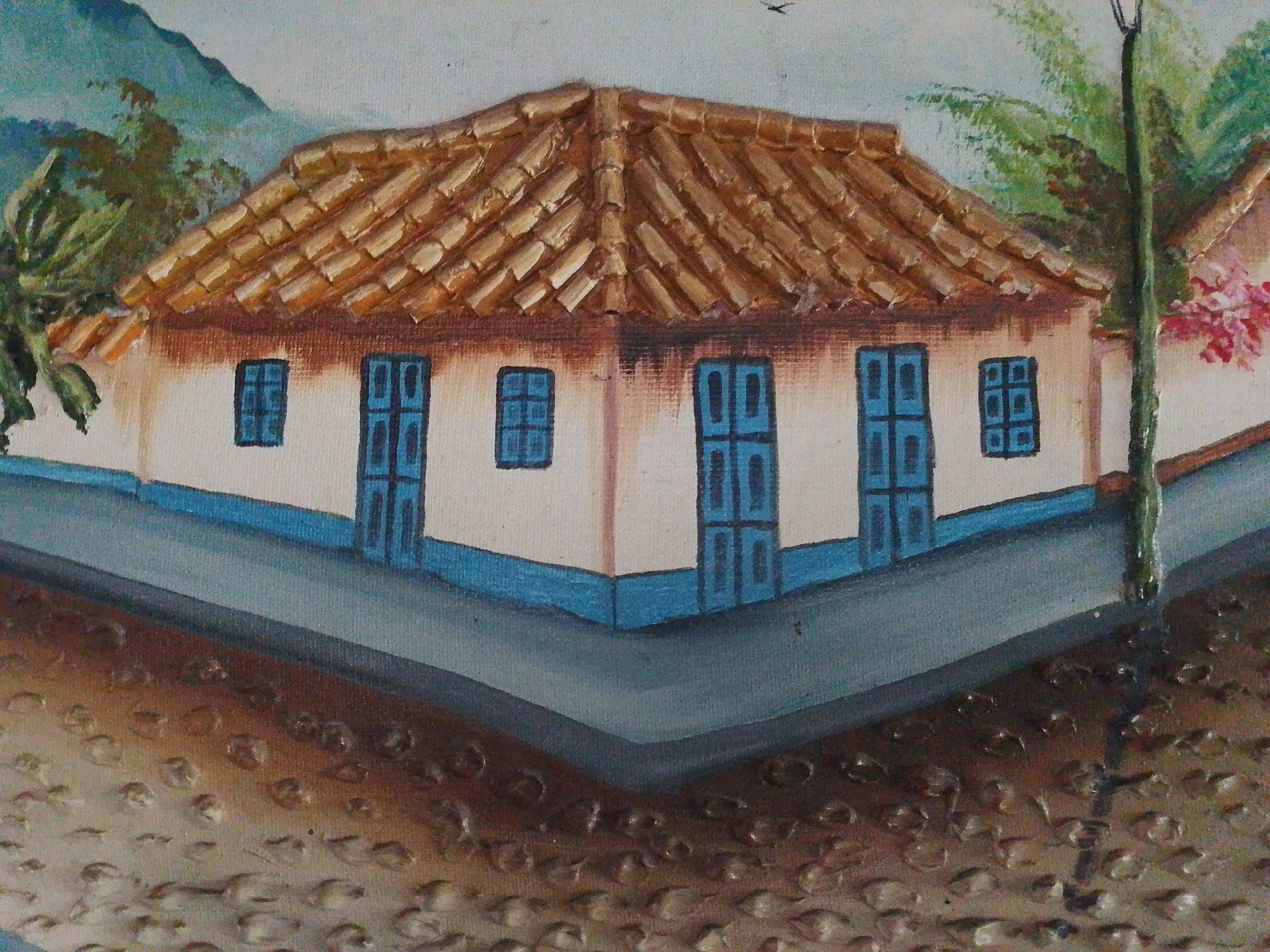 The image depicts a street corner in Tinaquillo, Venezuela, featuring a one-story building with a terracotta tile roof and blue-trimmed windows and doors. The building sits at a V-shaped intersection with a blue-gray road surface. The foreground shows a textured brown surface representing the ground. In the background, glimpses of greenery and mountains can be seen. A lamp post stands at the right side of the building.