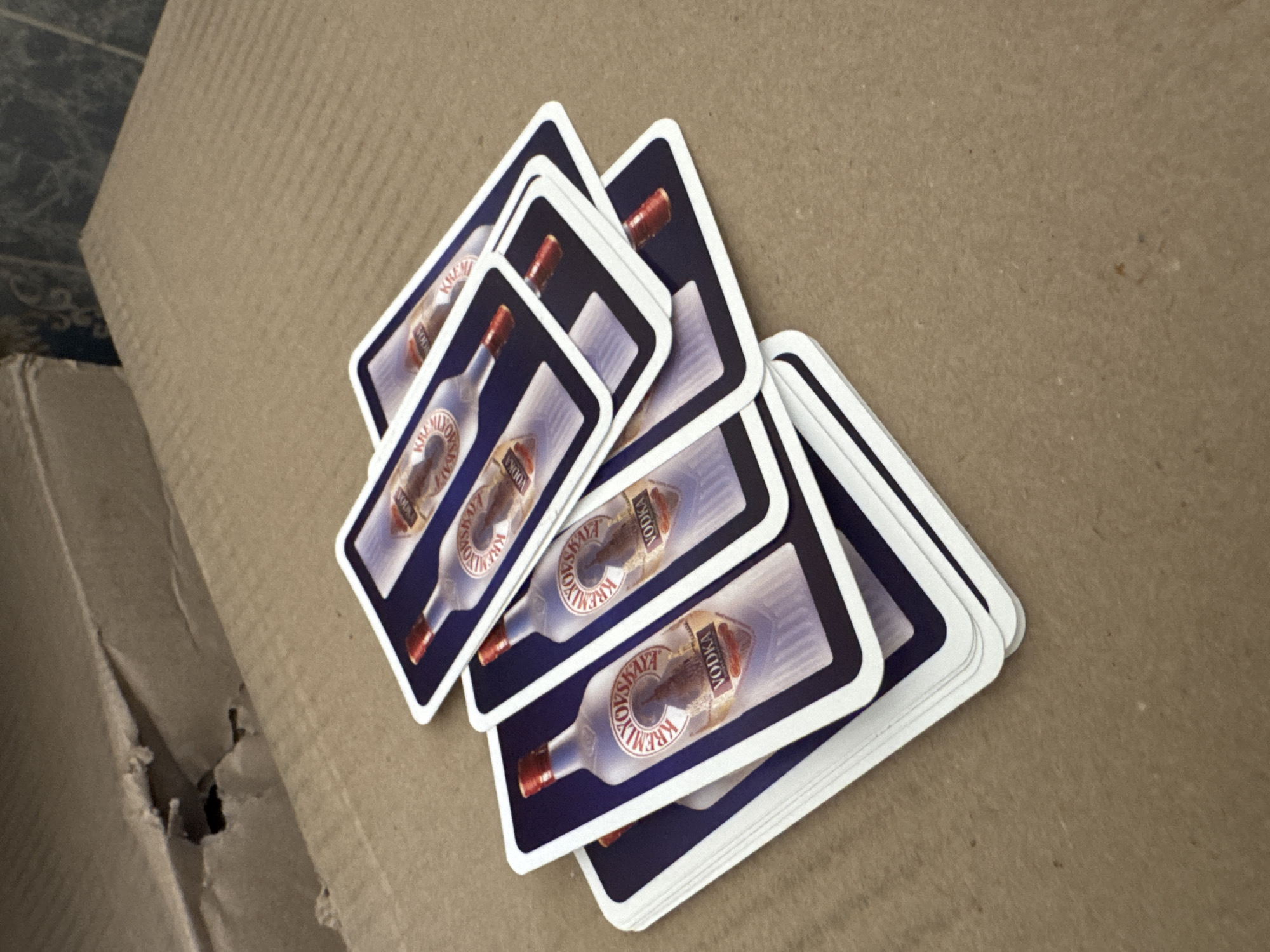 The image shows a pile of playing cards, each featuring an image of a bottle of "Kremlyovskaya" vodka. The cards are fanned out on a textured cardboard surface. The lighting suggests an indoor setting, likely daytime, with no strong shadows. The overall scene is casual and appears to be a close-up shot focusing on the cards. The cards have rounded corners and a dark blue border around the image. The visible text on each card is "VODKA" and "KREMLYOVSKAYA". The background is a plain, brown, corrugated cardboard surface, possibly part of a box. There are no people visible in the image. The location context provided is Rozdilna, Ukraine.