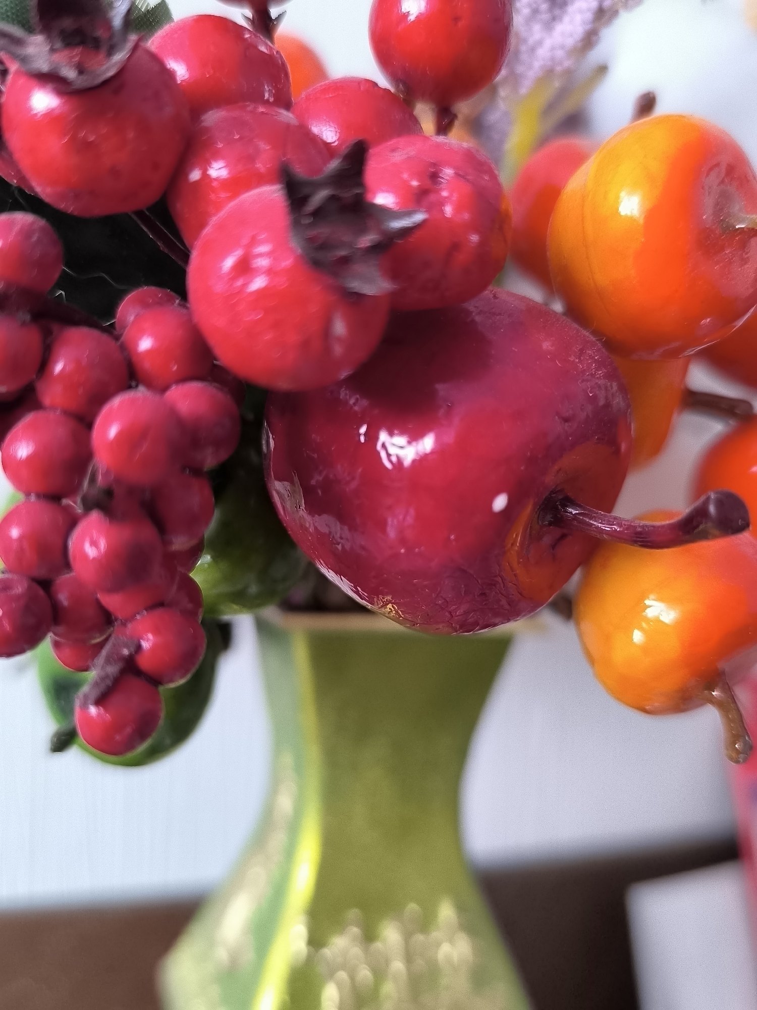 The image depicts a close-up of artificial berries and fruits arranged in a lime-green vase. The artificial fruits are varied in color, including bright red, deep red, and orange. Some of the red berries are clustered together, resembling a small bunch of grapes, while others are larger and rounder, possibly representing apples or persimmons. The orange fruits are also round and appear glossy. The vase has a curved shape, widening at the top and then narrowing towards the base, with a shiny finish. The background is blurred, with hints of white and possibly other decorative elements. There are no people or discernible actions taking place. The scene is a still life arrangement of decorative items. The image does not provide any information to suggest a location in Klaipėda, Lithuania.