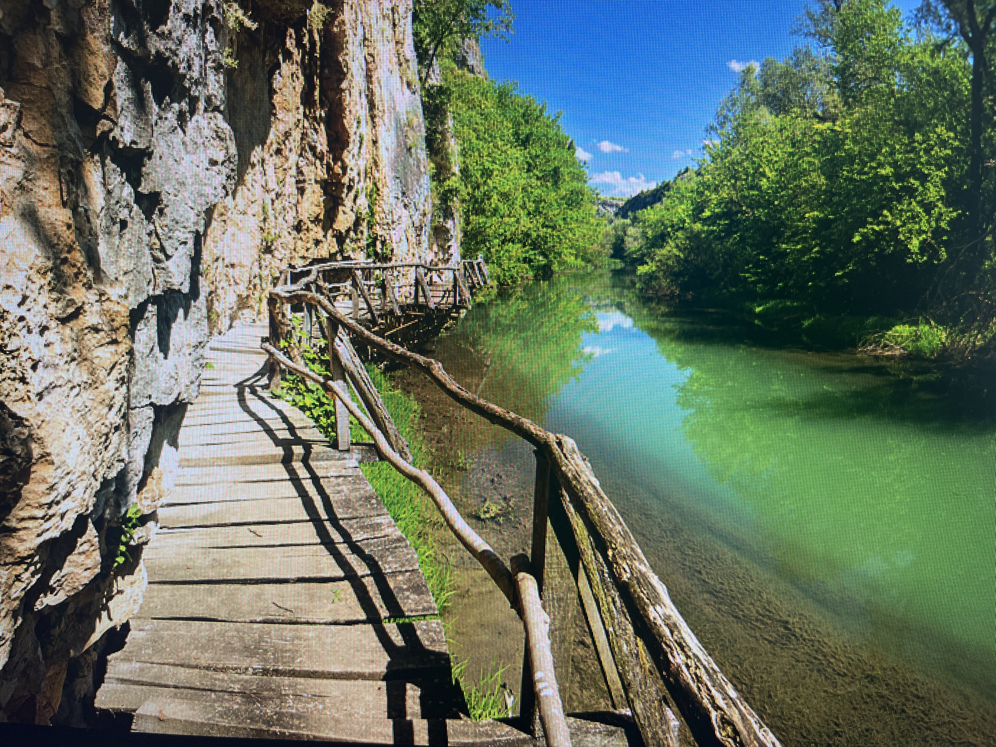 The image presents a vibrant natural landscape featuring a wooden boardwalk winding along a steep cliff face beside a tranquil river. The scene is captured in bright daylight under a clear blue sky dotted with a few wispy clouds.
On the left, a rugged, textured rock cliff rises prominently, serving as the support for a rustic wooden walkway. The walkway is constructed from horizontal planks and features an irregular, gnarled wooden handrail that follows the path's curve. Strong shadows from the railing and the cliff are cast across the walkway, indicating ample sunlight, likely suggesting late morning or early afternoon.
To the right, a calm river flows, its water exhibiting a striking green-blue coloration that mirrors the sky and surrounding foliage. The riverbed is visible through the clear water, revealing a textured, rocky bottom. The opposite bank is densely covered with lush, vibrant green trees and vegetation, creating a rich canopy that extends into the distance, hinting at a canyon or gorge.
No people are visible, emphasizing the untouched beauty and serenity of the location. The scene evokes a sense of peaceful exploration and natural wonder, characteristic of a scenic hiking trail. This beautiful natural setting is located in Bulgaria, with Varna provided as the geographical context. There is no visible text in the image.