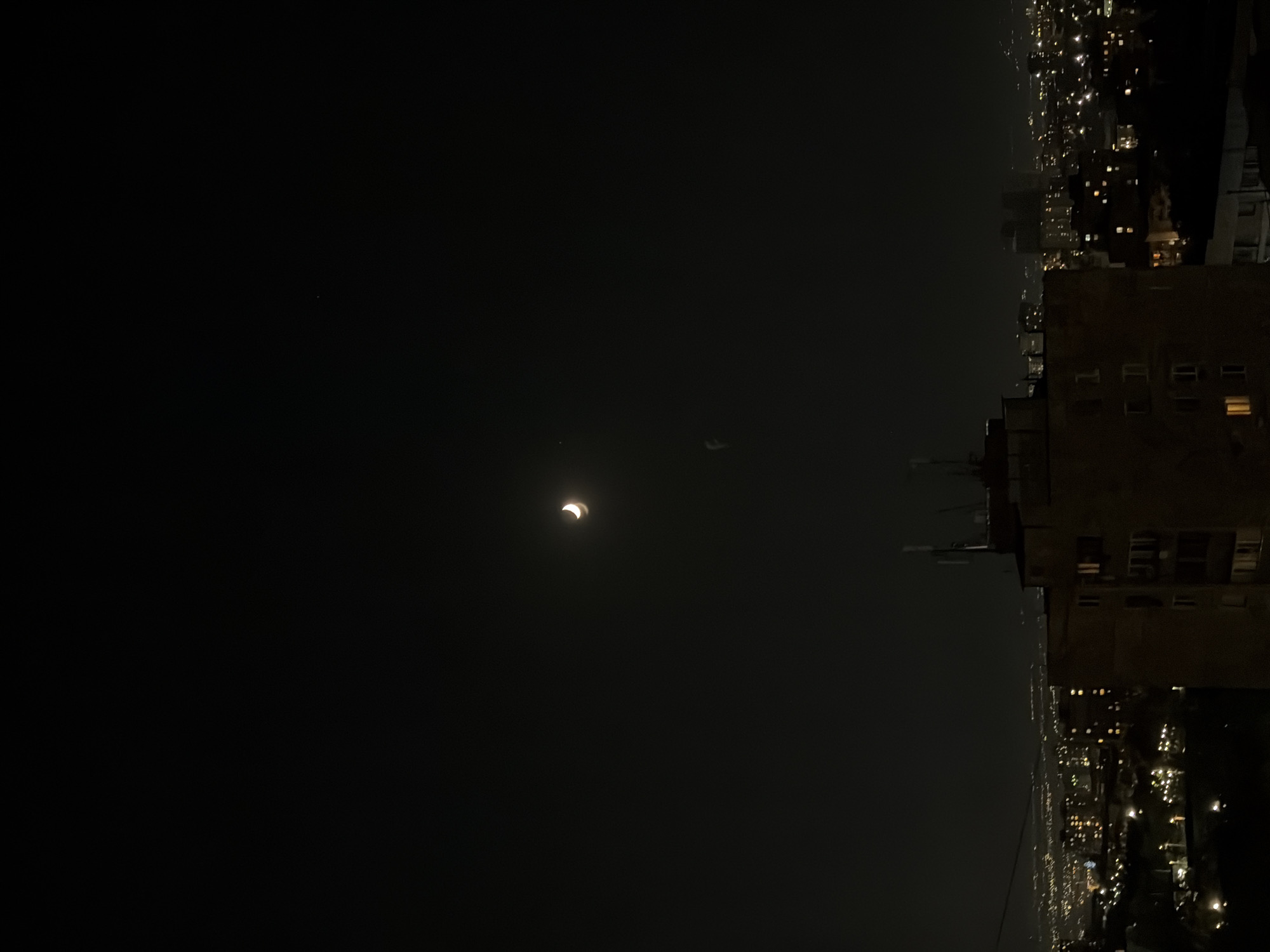 An image taken at night shows a crescent moon high in a dark sky above the city of Yerevan, Armenia. Below the moon, a small, faint object is visible, possibly a star or a distant balloon. The foreground is dominated by a dimly lit, multi-story building with visible windows. Beyond this building, the cityscape of Yerevan stretches out, illuminated by the lights of numerous buildings and streetlights, creating a hazy glow against the dark horizon. The overall scene captures a tranquil nighttime view of the city under a lunar sky.