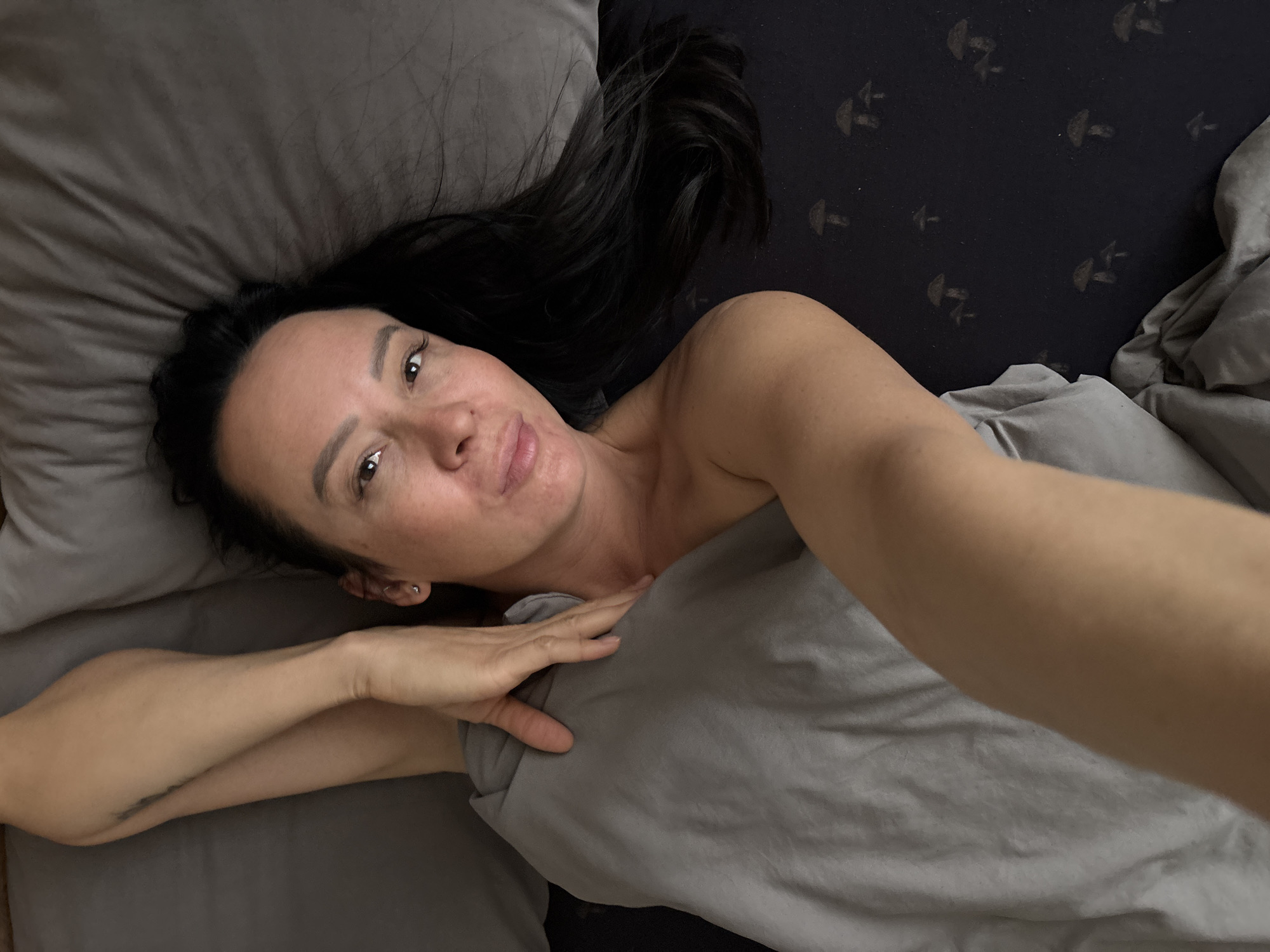 A woman is lying in bed, taking a selfie. She has long dark hair and is partially covered by a grey blanket. She is lying on grey bedding next to a darker, black patterned sheet or duvet. The location, Chaikovsky, Russia, cannot be confirmed from the image.