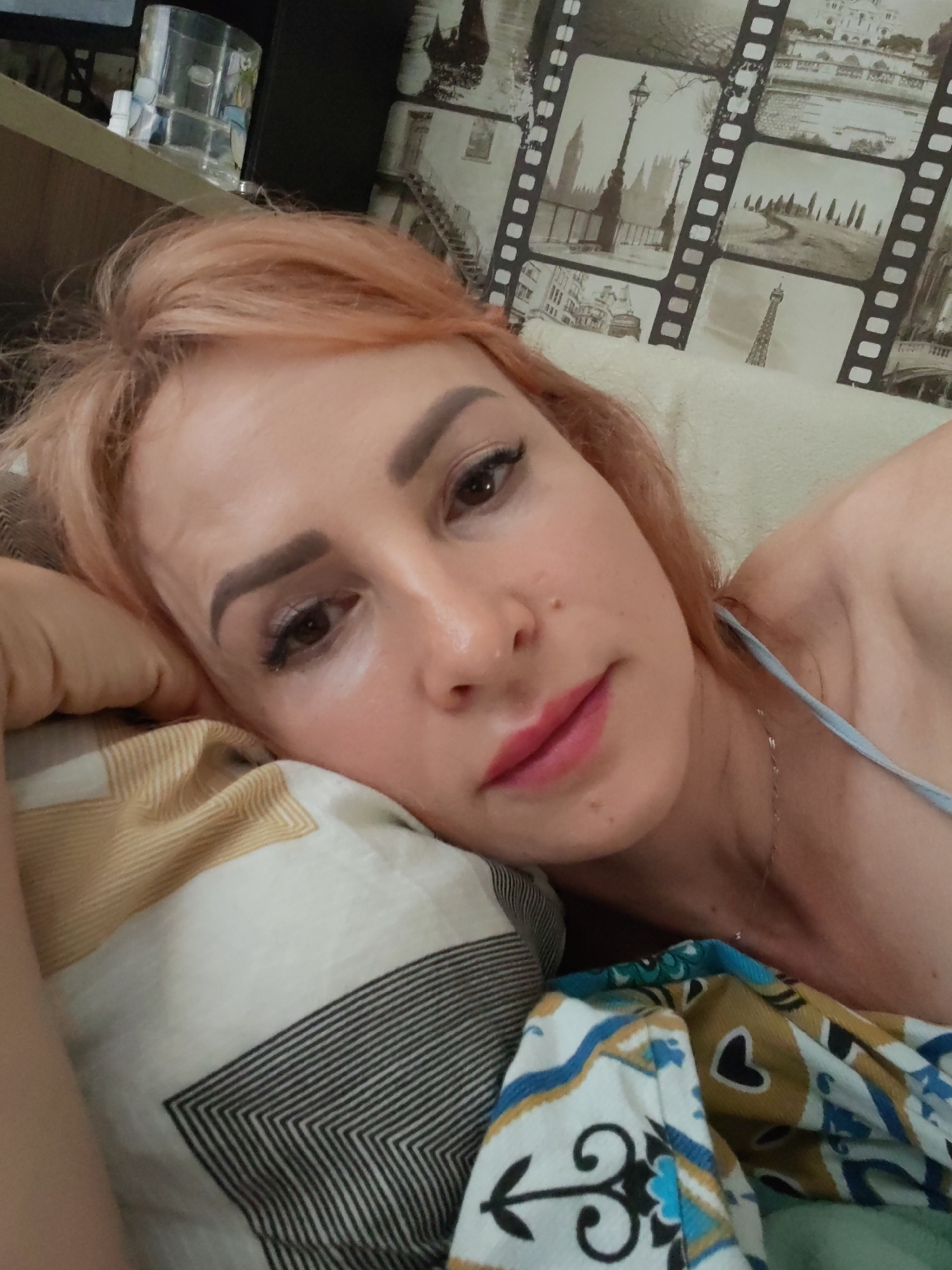 In a dimly lit indoor setting, a Caucasian woman with fair skin and peachy-blonde hair lies on her side, resting her head on a patterned pillow. She is looking directly at the camera, her brown eyes partially obscured by her eyelashes. Her lips are colored with pink lipstick. She wears a light blue strap visible on her shoulder, hinting at a possible top or dress. In the background, a wall is decorated with what appears to be a collage of black and white film strips featuring various city landmarks. The overall ambiance suggests a relaxed or intimate moment, with the woman appearing to be in bed or on a sofa. The image does not provide any specific contextual clues that would allow for its geolocation to Chaikovsky, Russia, or any other city.