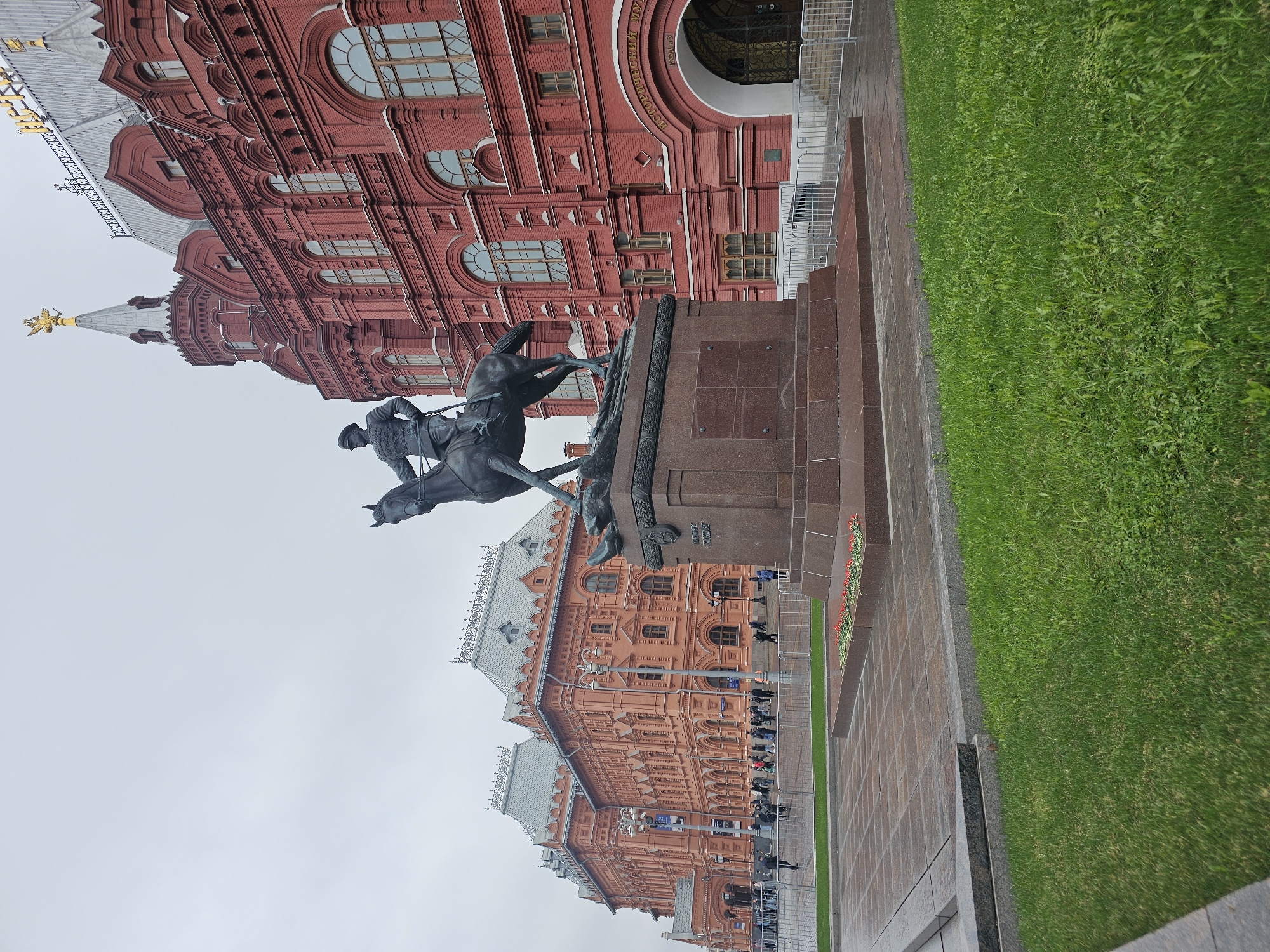 The image shows a statue of Kuzma Minin and Dmitry Pozharsky in front of the State Historical Museum on Red Square in Moscow, Russia. The statue is a bronze sculpture of the two figures on horseback, commemorating their leadership in the 17th-century Russian resistance against the Polish-Lithuanian Commonwealth. The State Historical Museum, a large red-brick building with ornate architectural details, serves as the backdrop. A line of people is near the entrance of the museum. There is a patch of green grass and paved ground in the foreground. The sky is overcast.
