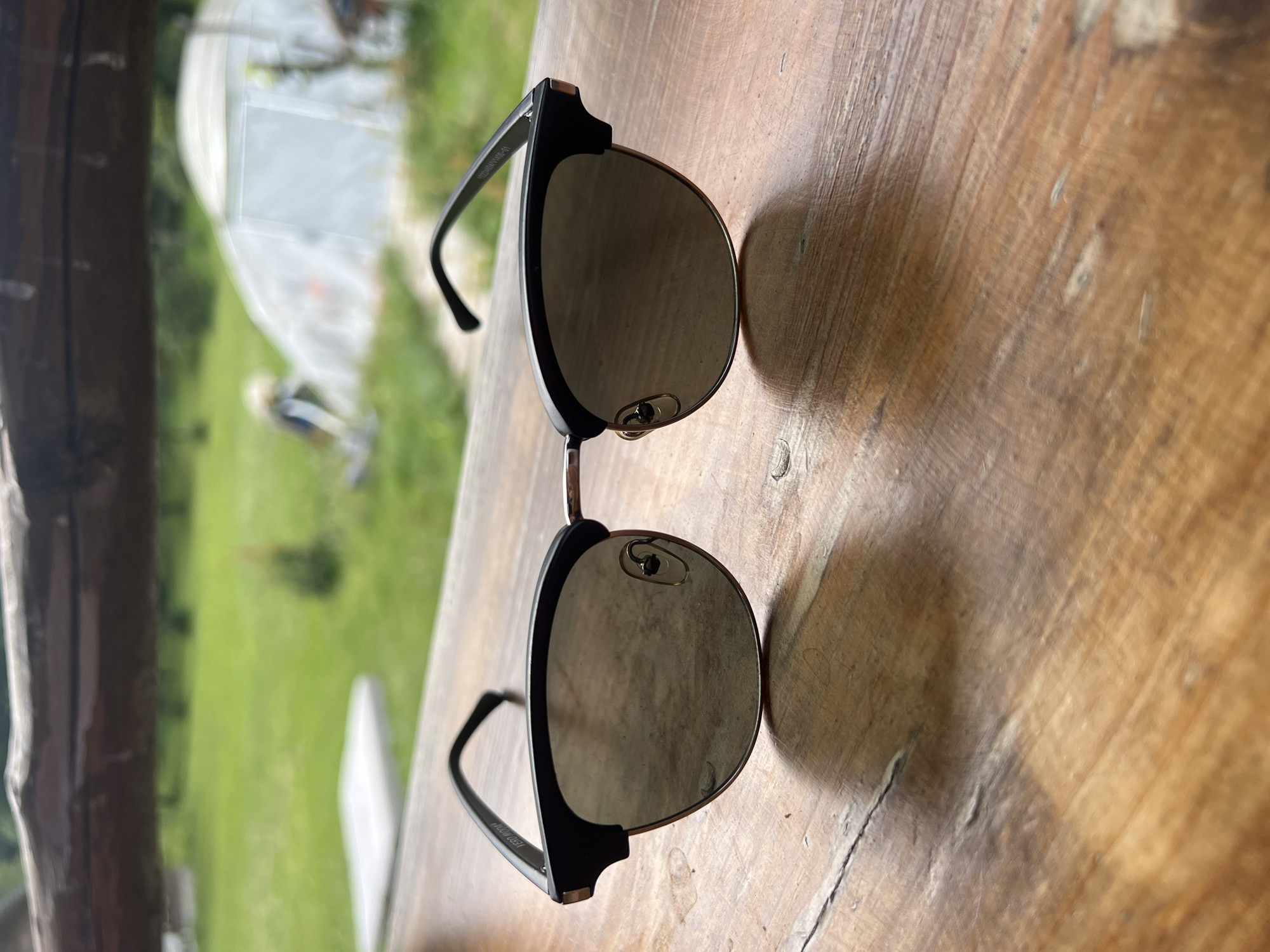 A pair of black and silver clubmaster-style sunglasses rests on a wooden table outdoors. The sunglasses have a thick black top rim and a metallic silver bottom rim. The lenses are tinted and reflective. The wooden table has a visible grain and appears weathered. In the background, a green grassy field stretches towards a white greenhouse-like structure. The overall scene suggests a casual outdoor setting in a rural or semi-rural environment, possibly in Norviliškės, Lithuania, given the context.