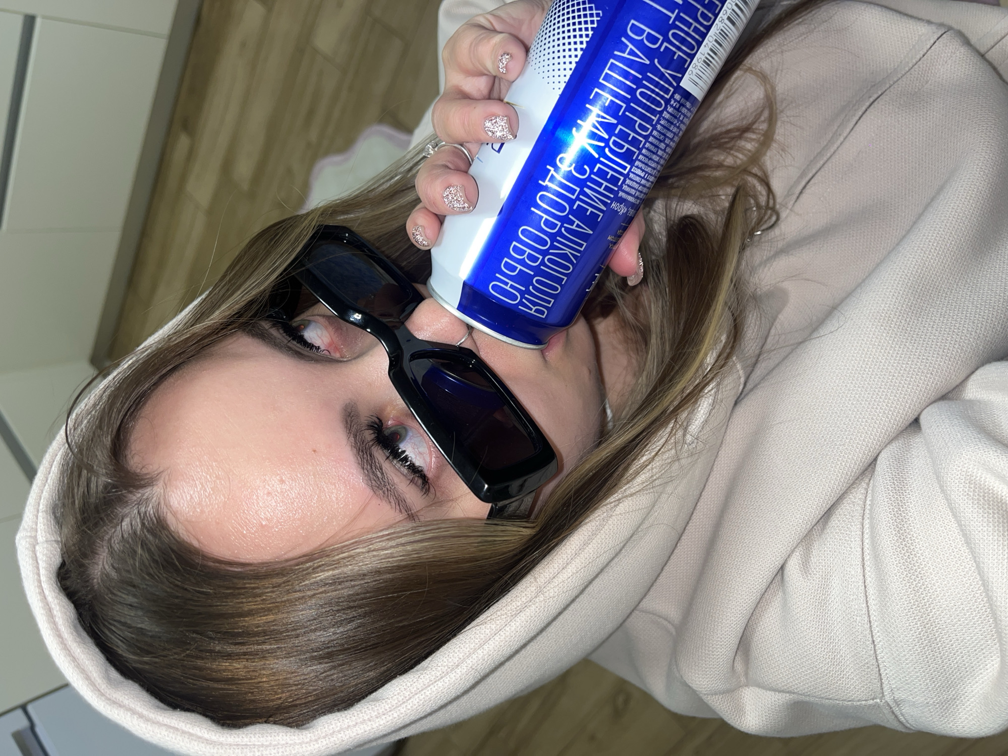 A young person, likely female, is featured in a tilted, close-up shot. She has long brown hair with lighter highlights, wears a light-colored hooded sweatshirt, and dark rectangular sunglasses that rest on her nose. Her eyes are visible above the sunglasses, with long, dark eyelashes. Her hand, adorned with rings and sparkly light-colored nail polish, holds a blue and white cylindrical can to her face, partially obscuring her mouth, suggesting she is drinking from it. She is looking towards the camera.
The background is an indistinct indoor setting with light-colored walls and a wooden floor, suggesting a casual home environment. The visible text on the can is in Russian, prominently displaying the warning: "ВРЕДНОЕ УПОТРЕБЛЕНИЕ АЛКОГОЛЯ ВАШЕМУ ЗДОРОВЬЮ" (Harmful consumption of alcohol to your health), confirming it is an alcoholic beverage. Other discernible Russian words on the can include "Крон" and "ПШЕНИЦА" (wheat). A barcode with the numbers "10682 04 1986" is also present. The overall mood is casual and relaxed. The location context for the image is Krasnodar, Russia, which is consistent with the Russian text on the object.