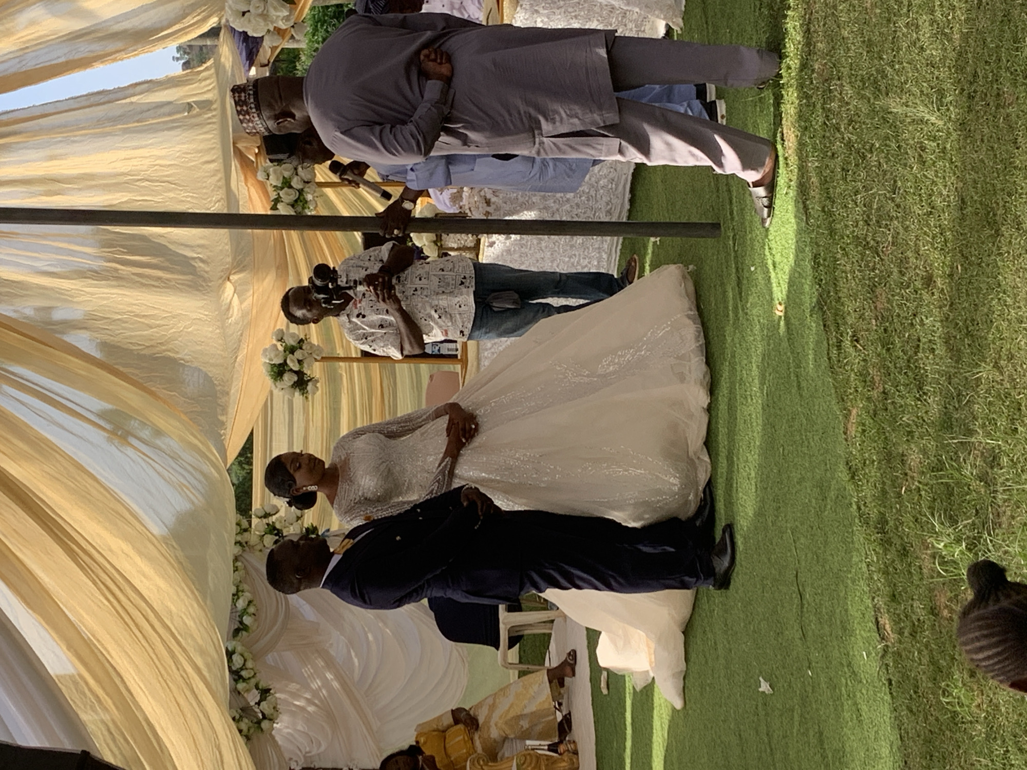 The image depicts a wedding scene likely in Jos, Nigeria, given the cultural attire of some guests. A bride and groom are centrally located, the bride in a long, shimmering white gown, and the groom in a dark suit. They are surrounded by guests, including a man in traditional Nigerian attire with a patterned hat, and another in a grey tunic. A photographer is also present, capturing the event. The wedding is held under a decorated tent, with cream-colored draped fabric and floral arrangements as decoration. The event takes place on a lawn area with green grass.