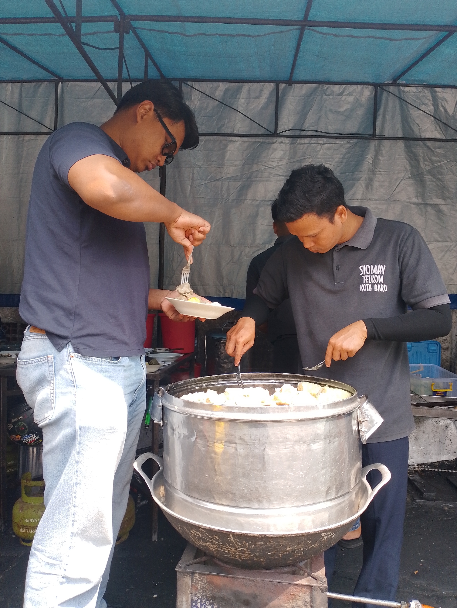 Two men are preparing siomay at a street food stall in Yogyakarta, Indonesia. The man on the left, wearing a navy t-shirt and light blue jeans, is holding a white plate with a fork in his right hand, scooping food from a steamer. He has sunglasses on. The man on the right, wearing a grey polo shirt with "SIOMAY TELKOM KOTA BARU" printed on it and black long sleeves underneath, is also using a fork and spoon to prepare food from the same large, aluminum steamer. The steamer is placed on a metal stand over a heat source, possibly a gas burner, indicated by a yellow gas cylinder visible to the left. The stall is covered by a light blue tarp supported by a metal frame. The background consists of a grey, possibly corrugated, material.
