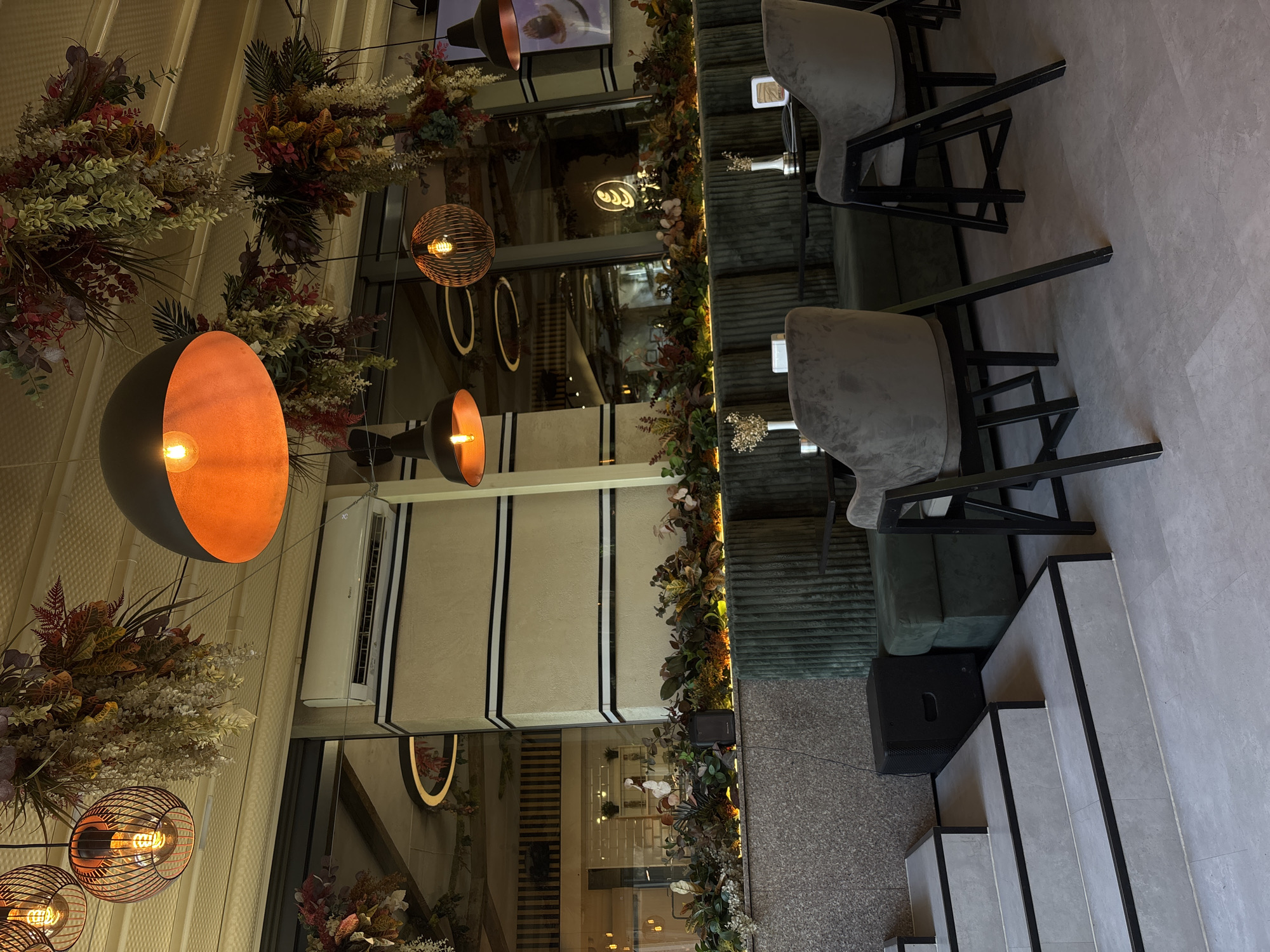 The image shows the interior of a modern cafe, likely located in Plovdiv, Bulgaria, with a focus on contemporary design and warm lighting. Hanging from the ceiling are several decorative light fixtures with exposed bulbs, including a large black dome-shaped pendant with an orange interior and intricate wire-framed spherical lamps. Floral and foliage arrangements, featuring a mix of autumnal colors, are suspended from the ceiling and draped along the upper walls, adding a touch of naturalistic decoration.
The cafe's seating area features plush, dark green ribbed banquettes and matching accent chairs with grey velvet upholstery and black metal frames. Small, dark tables are placed in front of the seating. A series of grey concrete steps with black trim leads up from the foreground. In the background, a long, textured wall with horizontal black stripes is visible, and an air conditioning unit is mounted above it. Large windows reflect the interior and exterior, revealing more of the cafe's ambiance. A black speaker sits on the floor near the base of the steps. The overall atmosphere is sophisticated and inviting, designed for a comfortable and stylish cafe experience.