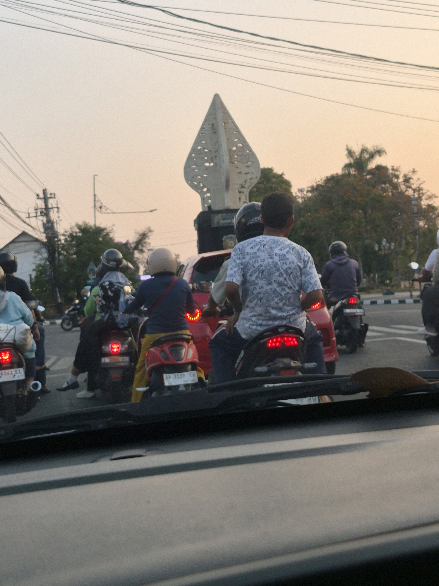 The image shows a traffic scene in the city of Purworejo, Indonesia, likely at dusk given the warm, hazy lighting and the clear sky. The foreground is dominated by the interior of a vehicle, with a dark dashboard and windshield. In the midground, a group of motorcycles and their riders are stopped in traffic. Several riders are wearing helmets, and one rider in the center is wearing a white t-shirt with a patterned design. The motorcycles are varied in color and style. In the background, a distinctive, tall, white modern sculpture with a pointed top and cutouts is visible, a landmark that helps identify the location as Purworejo. Trees and power lines are also present, suggesting an urban or semi-urban environment. The overall impression is one of a typical evening traffic jam in an Indonesian city.