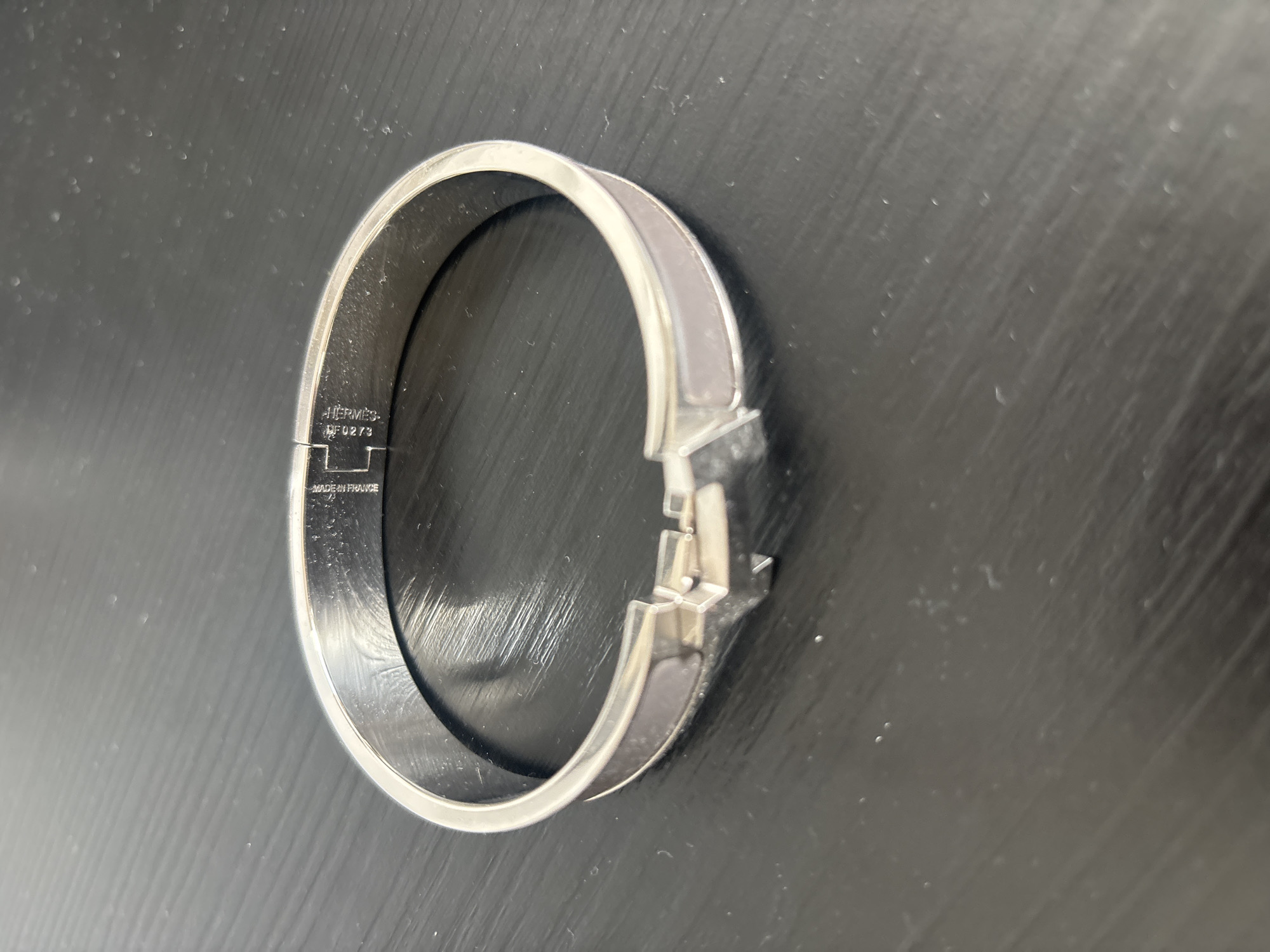 A silver-toned Hermès bracelet with a light-colored inlay is displayed on a dark, textured surface, likely a table. The bracelet is a cuff style with a hinge closure. Engraved markings are visible on the inner band of the bracelet. The scene is lit from above and to the left, casting subtle shadows. The overall setting suggests a controlled environment, possibly a retail display or a personal collection. There is no indication of a specific location within Dubai, United Arab Emirates.