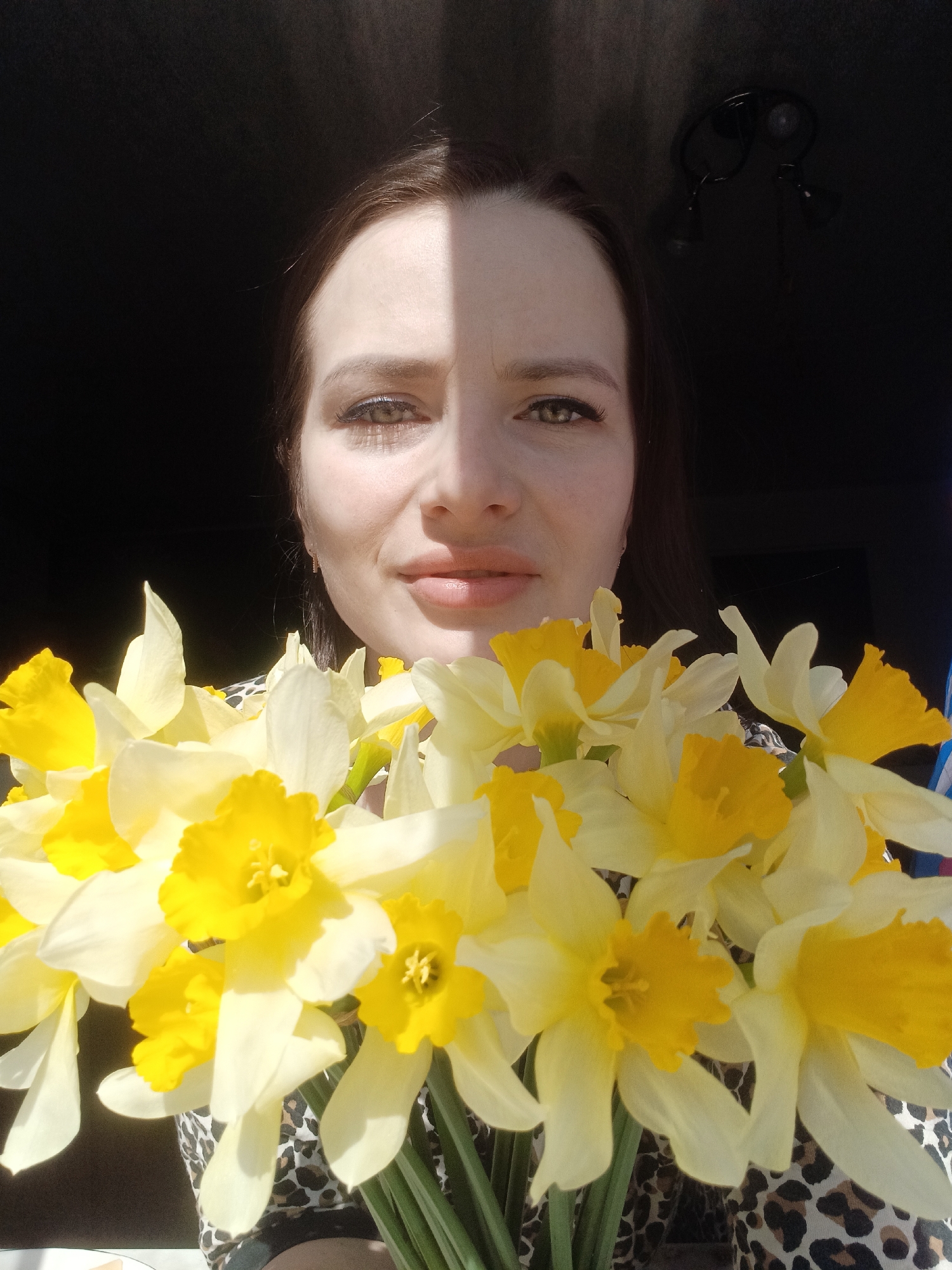 An adult woman is pictured holding a vibrant bouquet of yellow and white daffodils. She looks directly at the viewer with a subtle, closed-lip smile. Her face is partially illuminated by a strong, direct light source, casting a distinct shadow down the center of her face, with the left side (from the viewer's perspective) appearing darker. Her dark hair frames her face, and her eyes are clearly visible.
The daffodils dominate the lower portion of the image, showcasing their bright yellow trumpet-shaped coronas against white or pale yellow petals. The flowers appear fresh and in full bloom. The woman is wearing a garment with a brown and black leopard print pattern, visible behind the flowers.
The background is predominantly dark, suggesting an indoor setting with a strong natural light source, likely a window, positioned to her right. In the upper right corner, a dark, ceiling-mounted light fixture with two adjustable spotlight-like heads can be faintly discerned against the dark wall or ceiling. The strong contrast between the sunlit areas and the dark background creates a dramatic effect.
The scene conveys a sense of cheerfulness and warmth, likely taking place during the daytime in sunny weather, in Rusalevka, Russia. There is no visible text in the image.