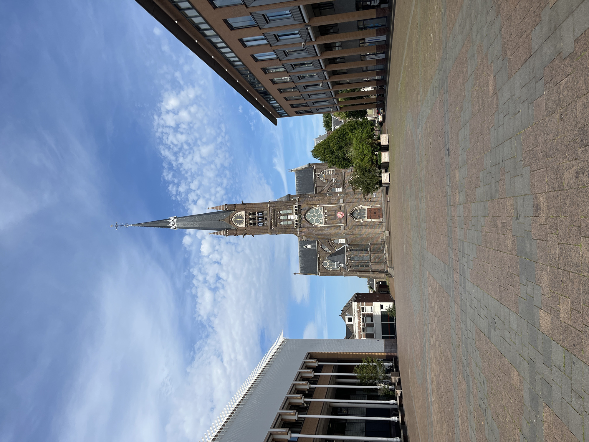 This vertical, full shot depicts an outdoor scene dominated by architecture and a bright blue sky with scattered white clouds. The foreground is a large, paved plaza with a pattern of reddish-brown and gray rectangular pavers. On the left side of the plaza, a modern building with a white facade and a series of vertical white columns is visible. Further back, a historical church with a tall, pointed spire and ornate stone detailing rises against the sky. The church features large arched windows, some with stained glass, and a clock face. Trees with lush green foliage are interspersed between the buildings. Another multi-story building with a brick facade and numerous windows is visible to the left of the church. The lighting suggests it is daytime, possibly late morning or early afternoon, with clear weather. There are no people or animals visible in the frame. The overall impression is one of urban architecture, with a contrast between modern and historical styles.