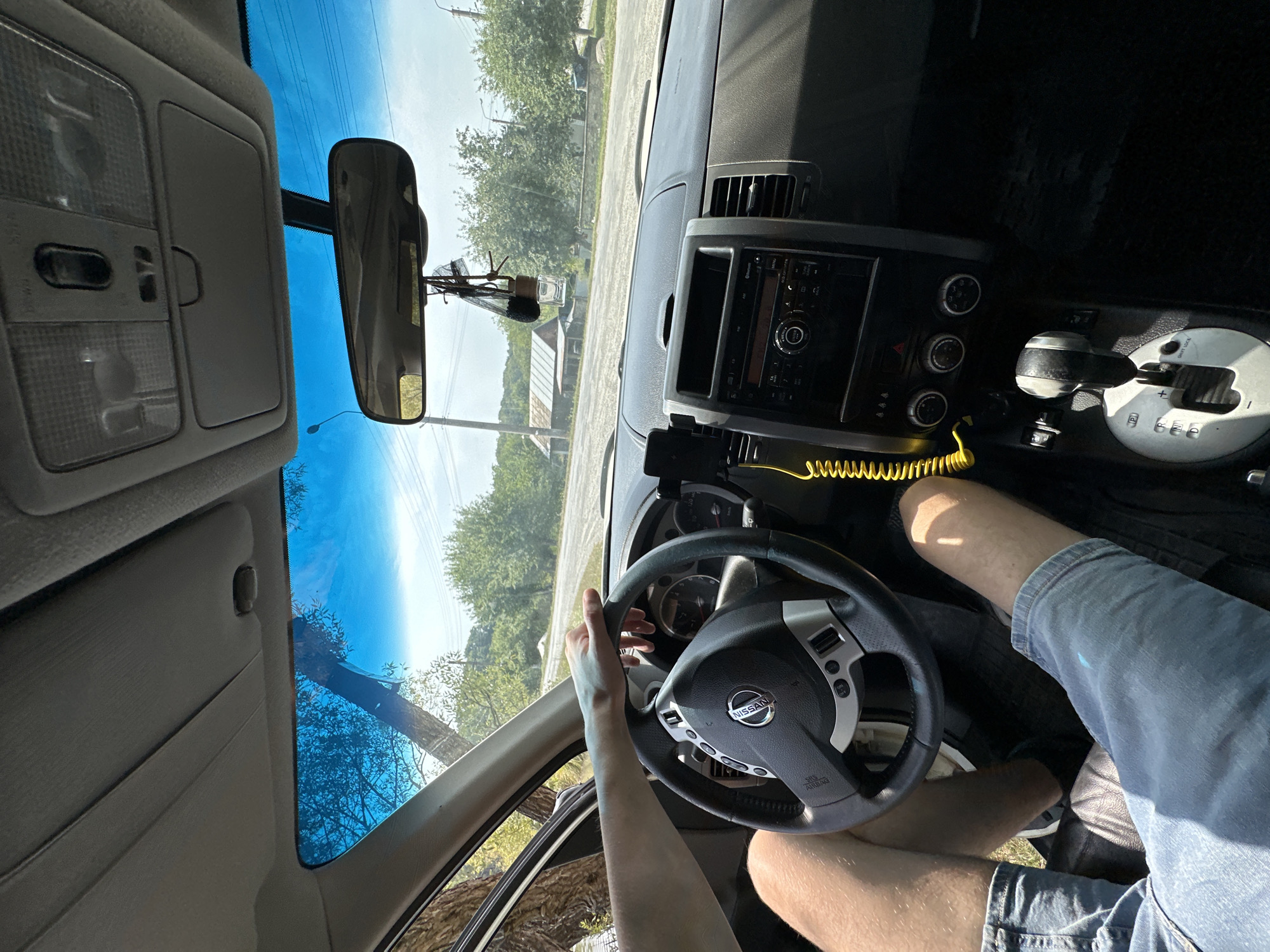 The image shows the interior of a car from the passenger's perspective, looking towards the driver's side. A person's arm and hand are resting on the steering wheel, and their legs are visible in the lower part of the frame, wearing blue jean shorts. The car's dashboard features a radio, air vents, and a gear shifter. A yellow coiled cable is attached to a phone mount on the dashboard. Through the windshield, a rural landscape is visible with trees, a road, and some buildings. The sky is bright blue with some clouds. This scene is located in the city of Makiv, Ukraine.