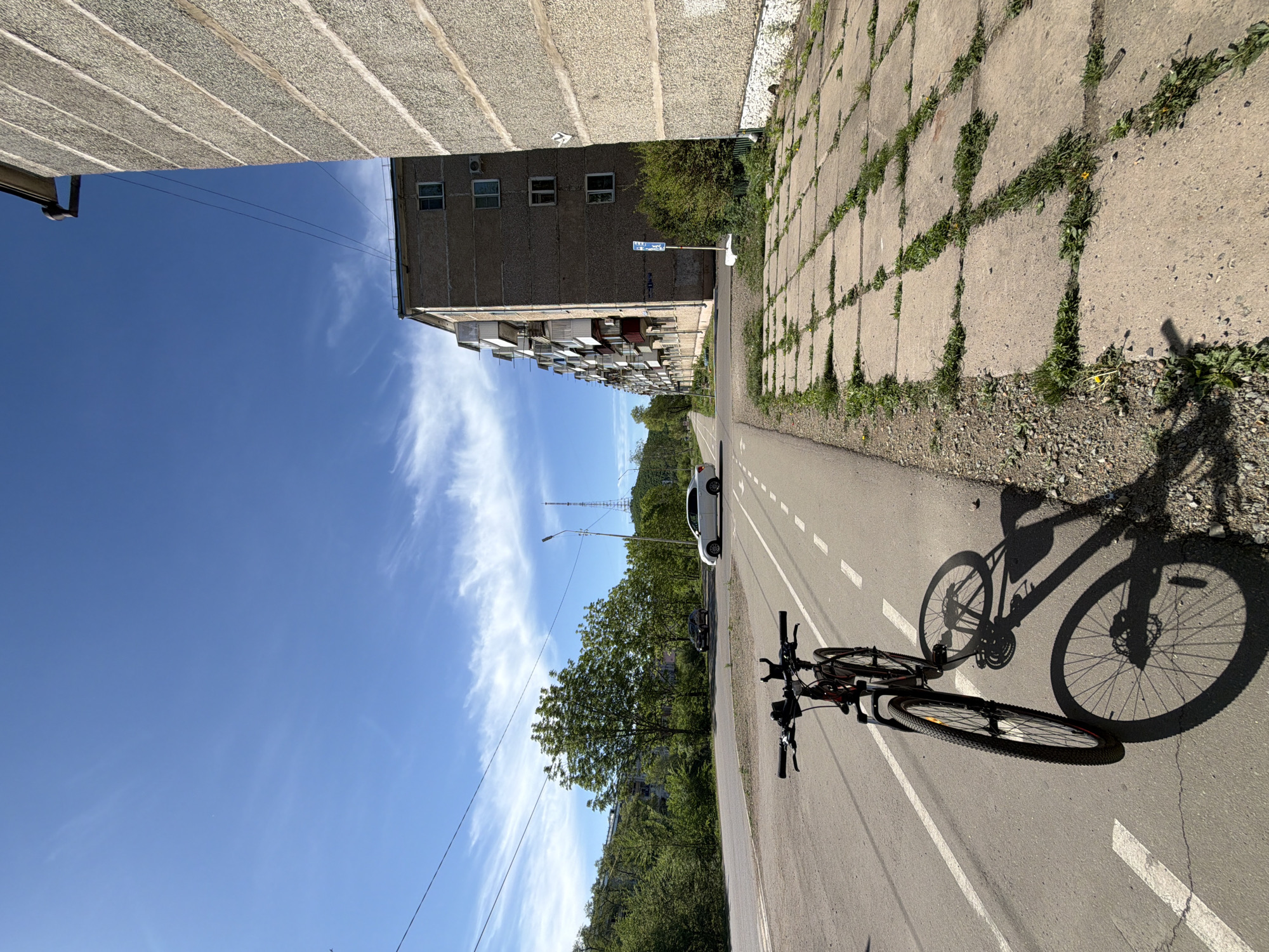 The image presents a street scene in what appears to be a residential area, likely located in Birobidzhan, Russia. The view is oriented along a paved path or roadway marked with dashed lines. A bicycle, casting a prominent shadow, lies along the path. A multi-story concrete apartment building stands to the left. Across the path, a concrete wall composed of large square blocks, interspersed with vegetation, can be seen. A few vehicles are parked further down the road. The sky is blue with light wispy clouds. The angle of the sun suggests it is either morning or late afternoon.