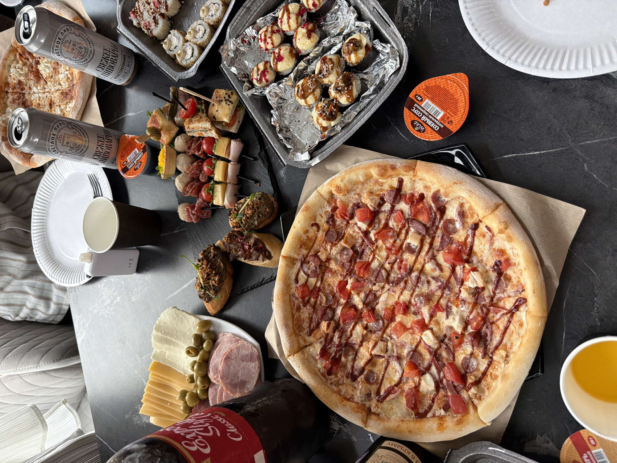 This is a top-down, close-up shot of a spread of food and drinks on a dark, textured surface, likely a table. The scene suggests a casual gathering or meal.
Dominating the frame is a large pizza, topped with cheese, sliced sausages, and diced tomatoes, drizzled with a dark sauce. To the left of the pizza, two silver cans of beer are visible, one slightly overlapping the other. The cans have Cyrillic text, with one clearly reading "СВЕТЛОЕ ПИВО" (Light Beer) and "СВЕЖЕЕ ПИВОВАРНИ" (Fresh Brewery).
In the upper left quadrant, a tray of sushi rolls, some topped with sesame seeds and drizzled with sauce, is presented. Adjacent to this, a platter holds an assortment of appetizers on skewers, including mini sandwiches, cherry tomatoes, cheese cubes, and small meatballs or falafel. Another dish appears to contain bruschetta-like appetizers with dark toppings.
A plate with sliced cheeses, ham, and olives is positioned in the lower left corner, next to a dark bottle with a red and gold label that includes the word "Classic" in English. A small, orange sauce container with Cyrillic text, reading "СОЕВЫЙ СОУС" (Soy Sauce), is near the pizza. A white paper cup and a white paper plate are also present.
The lighting suggests it could be indoors or a dimly lit outdoor setting. The food appears to be ready to be consumed, indicating a meal or party in progress. The presence of both pizza and sushi suggests a varied palate or a buffet-style offering. The overall impression is one of abundance and a relaxed, communal dining experience.