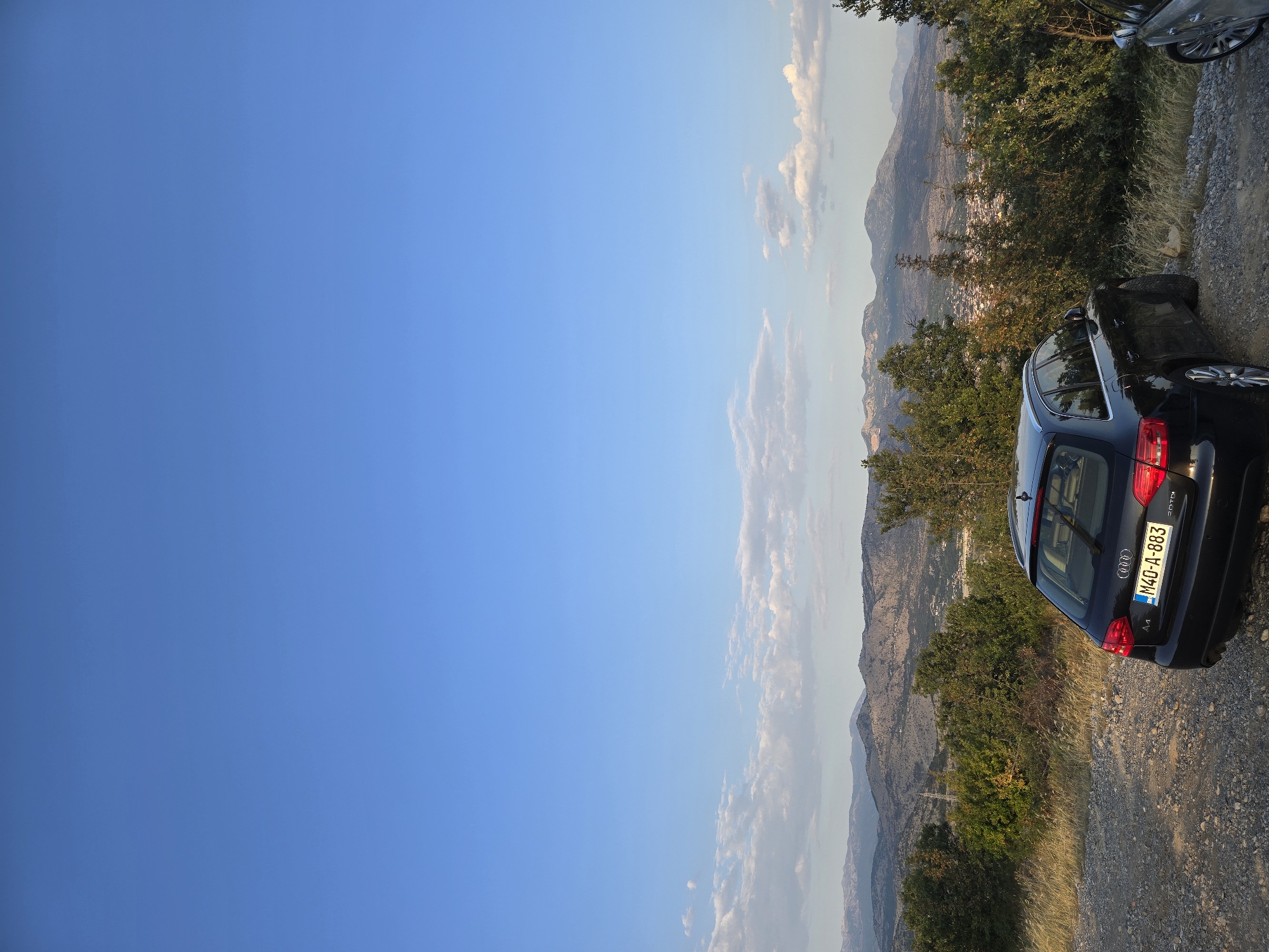 A black Audi A4 Avant station wagon is parked on a gravelly road overlooking a valley with a town and mountains in the background. The car has a license plate that reads "M40-A-883". The sky is clear and blue with some scattered clouds. The sun is setting, casting a warm glow on the landscape. The location is likely a viewpoint on the outskirts of Podgorica, Montenegro, offering a panoramic view of the city and the surrounding Dinaric Alps. The vegetation consists of green trees and dry grass. The overall atmosphere is peaceful and scenic.