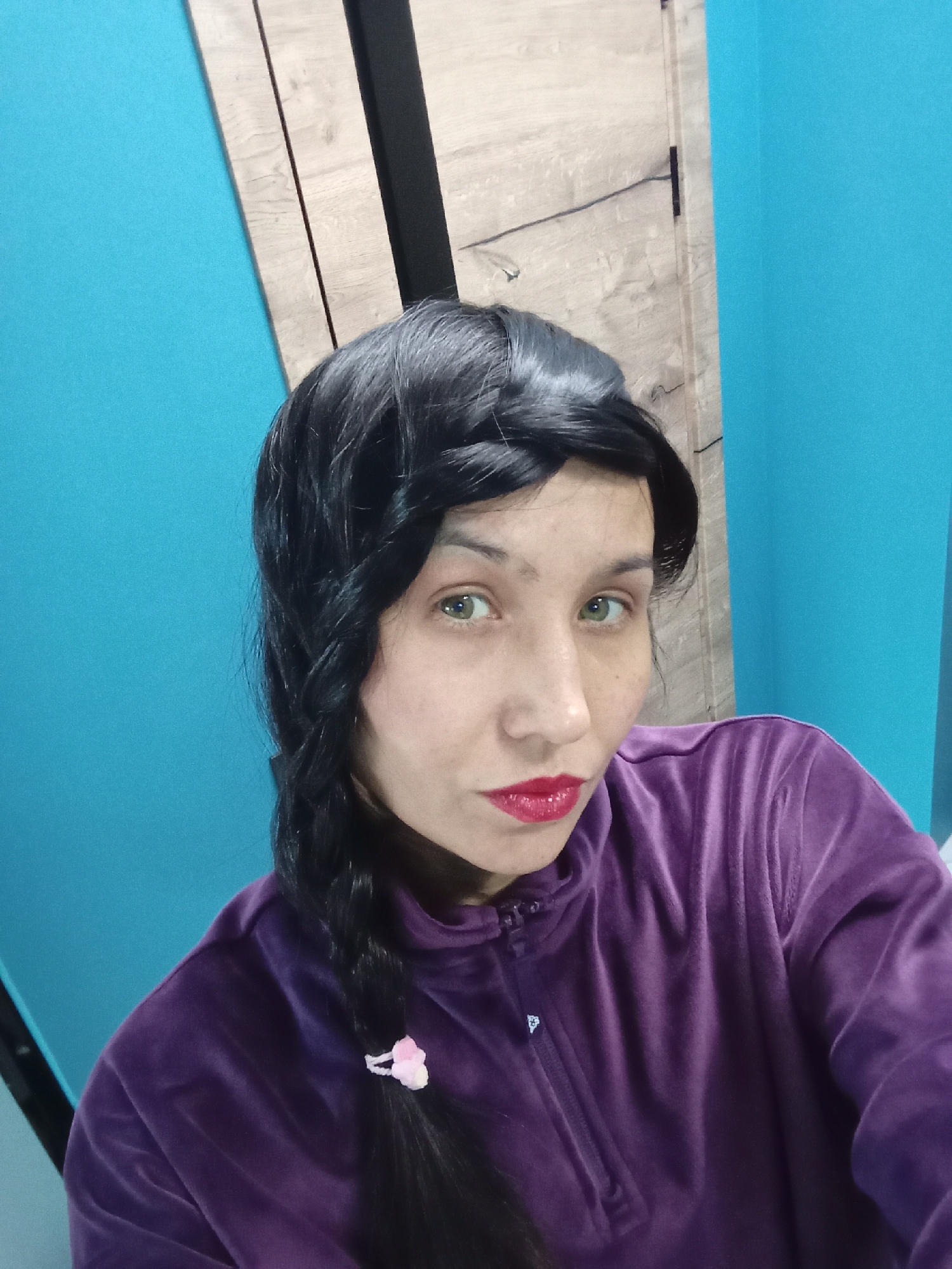 A woman with black hair braided to the left side, featuring a pink hair tie at the braid's end, is wearing a purple velvet zip-up hoodie. She is looking towards the camera, has light skin, green eyes, and red lipstick. The background consists of a blue wall, with a strip of wood paneling and a narrow black vertical structure visible behind her. The location is unknown but not specifically verified to be the city of Pervomaysky, Russia.