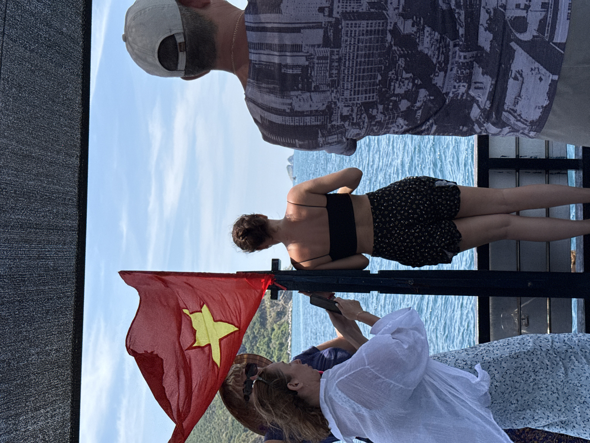 On a boat in Nha Trang Ward, Vietnam, a group of people are standing. A red Vietnamese flag with a yellow star is positioned on the left. A woman in a white top is holding a phone, possibly taking a picture. Another woman, wearing a black top and a patterned skirt, is resting her arms on a metal railing. A man with a beard and a baseball cap is facing away from the camera, wearing a patterned shirt. The blue sea and sky are visible in the background.