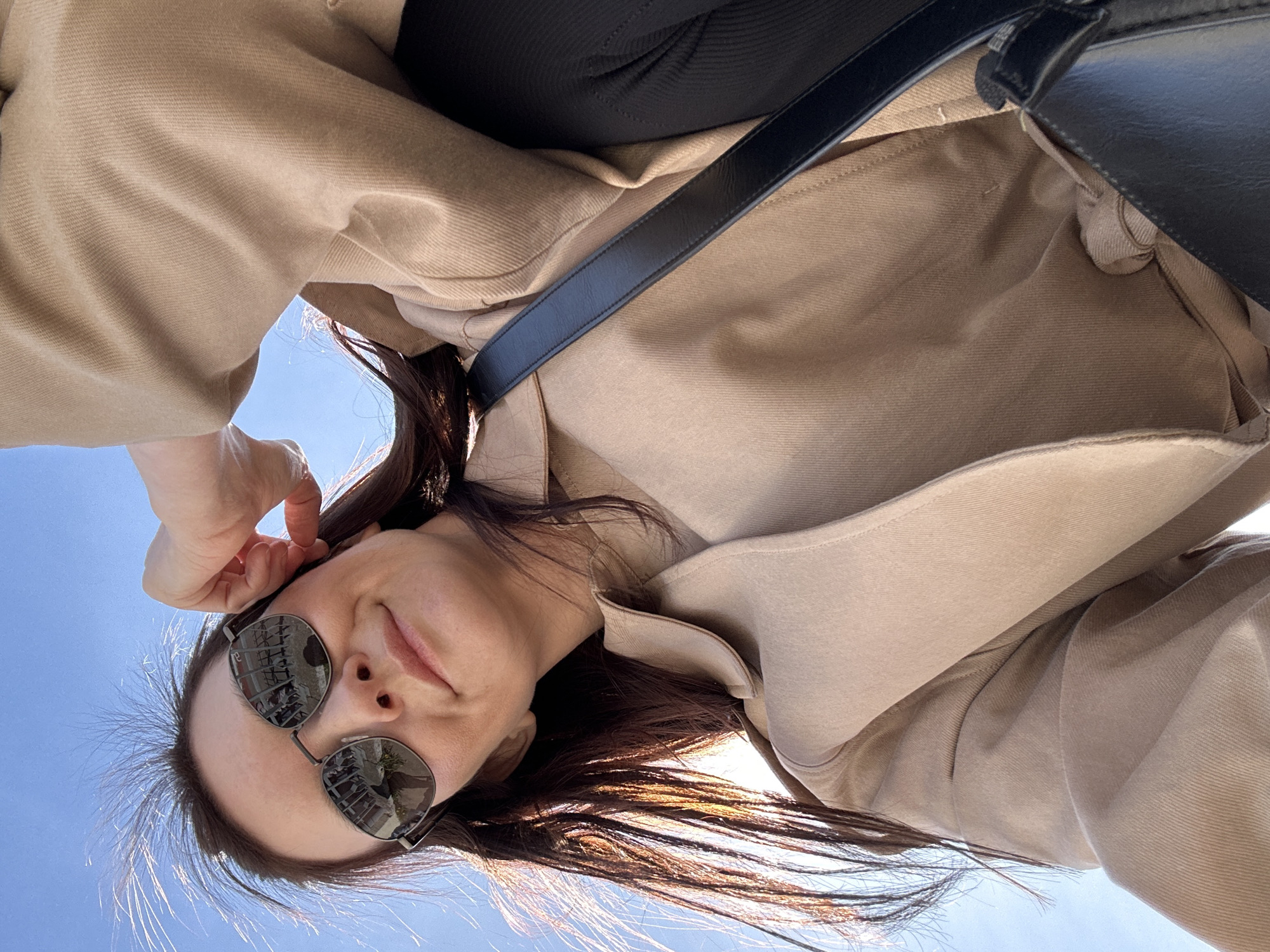 A woman wearing sunglasses and a beige coat is looking down at the camera from a low angle against a clear blue sky. She has dark hair and is holding her left hand near her head. A black bag with a strap is partially visible over her shoulder. The reflection in her sunglasses shows a blurred urban environment, potentially indicating the location is in a city. The city is Chaikovsky, Russia.