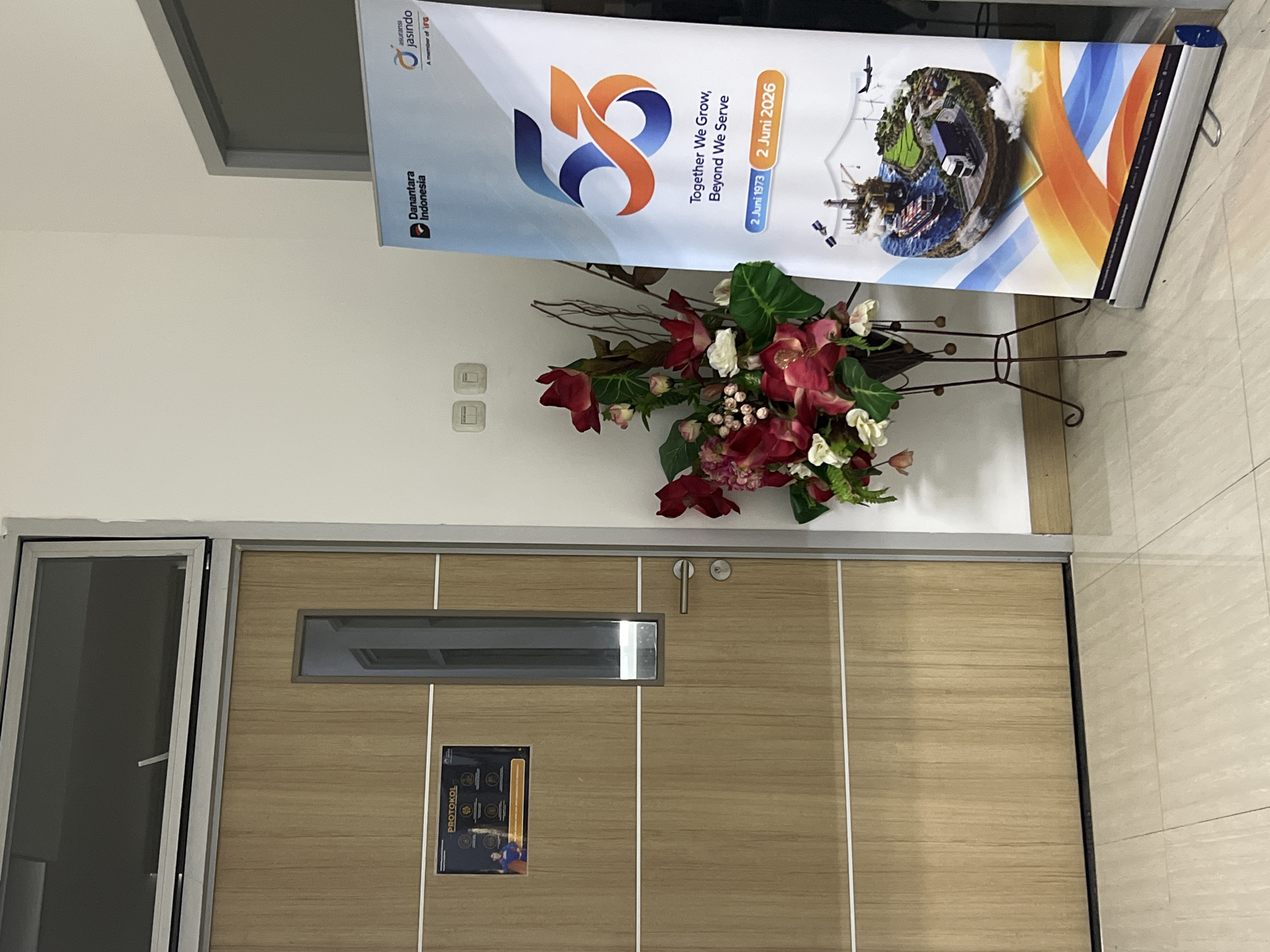 The image shows the interior of an office hallway. On the left, there is a wooden door with a rectangular window. A sign with the word "PROTOKOL" is attached to the door. To the right of the door, there is a bouquet of artificial flowers in a dark metal stand. In front of the flowers, a banner is displayed. The banner is white and features a large "53" graphic, along with text that reads "Together We Grow, Beyond We Serve" and "2 Juni 1973 - 2 Juni 2026". The banner also has logos for "Danantara Indonesia" and "asuransi Jasindo". The floor is tiled with light-colored ceramic tiles. Two light switches are visible on the wall behind the flower arrangement. The overall scene suggests an office environment, possibly celebrating an anniversary.