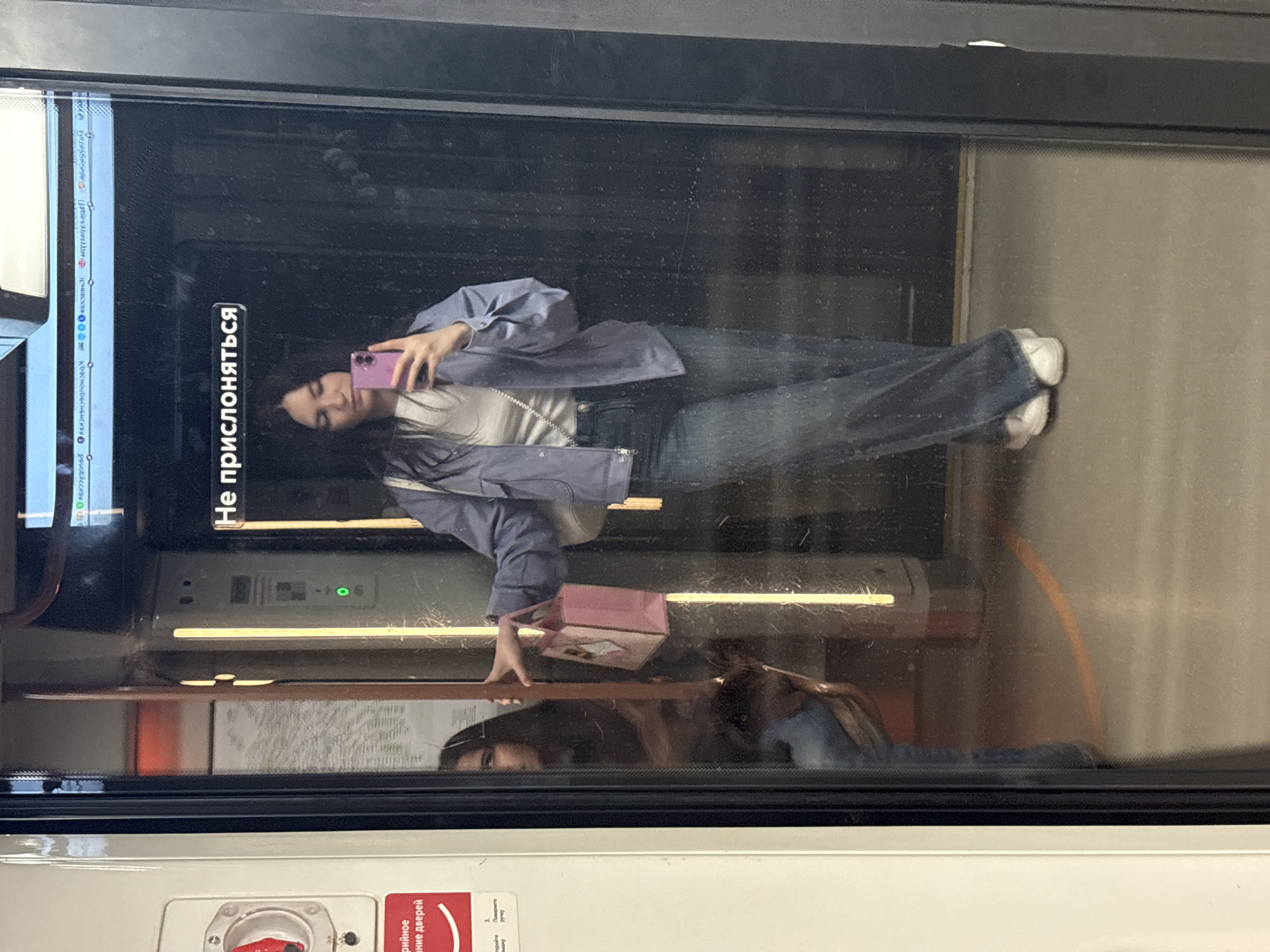 The image shows a young woman reflected in the glass door of a Moscow metro train. She is holding a pink smartphone in her hand, taking a selfie. She is wearing a light blue jacket, a white top, and blue jeans. She also has a pink bag.
The reflection shows the interior of the train, with signs in Russian. One sign reads "Не прислоняться," which means "Do not lean." There is also a diagram of the metro system visible in the door. The train appears to be in motion.