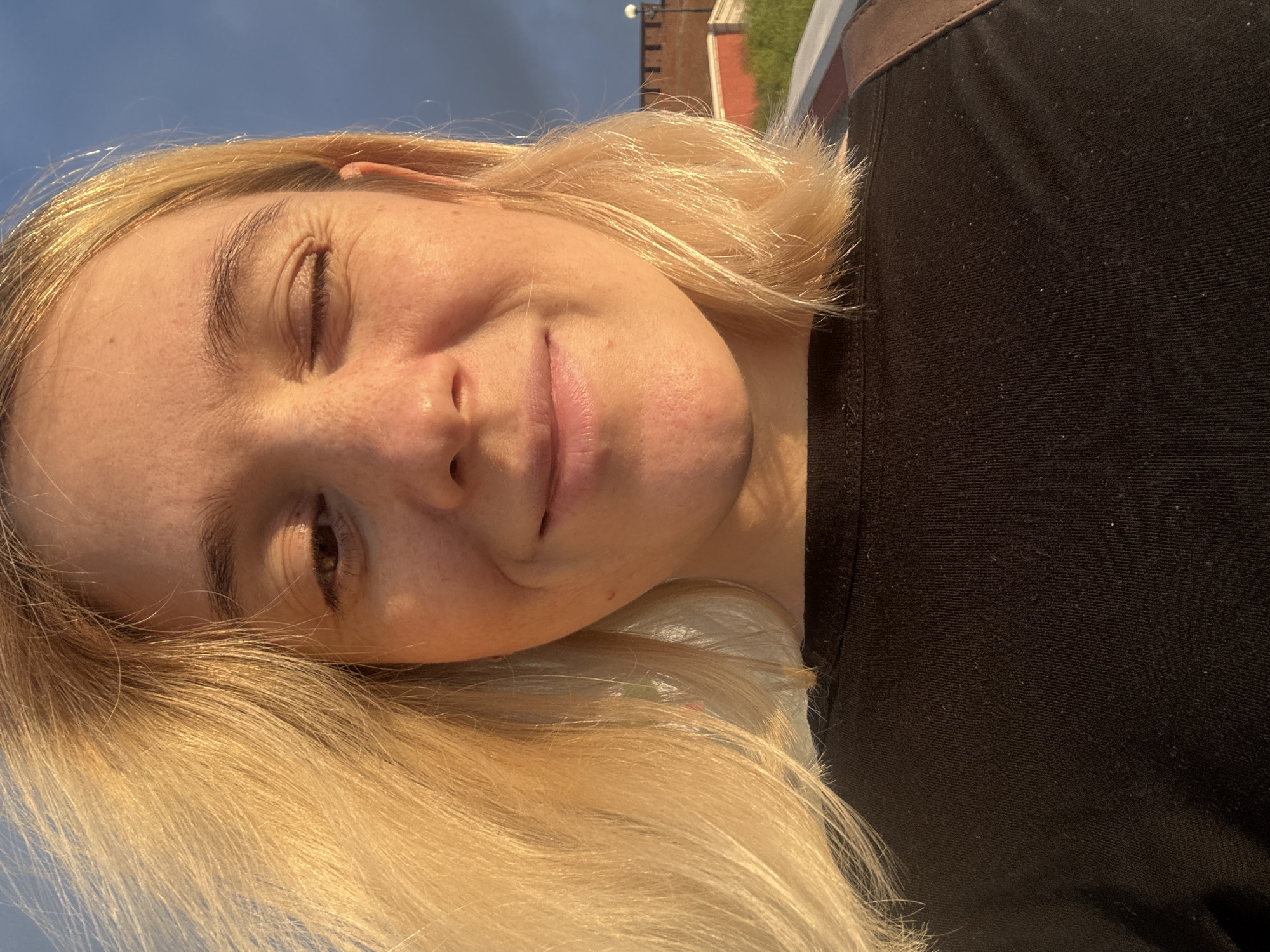 A woman with blonde hair and fair skin is taking a selfie outdoors. She is wearing a black t-shirt. Her right eye is closed in a wink, and her mouth is slightly open in a smile. Freckles are visible on her cheeks. The background shows a cloudy blue sky and a brick building with a lamppost, suggesting an urban setting. The lighting indicates it is either sunrise or sunset. The scene is likely in Nizhny Novgorod, Russia, given the provided context.