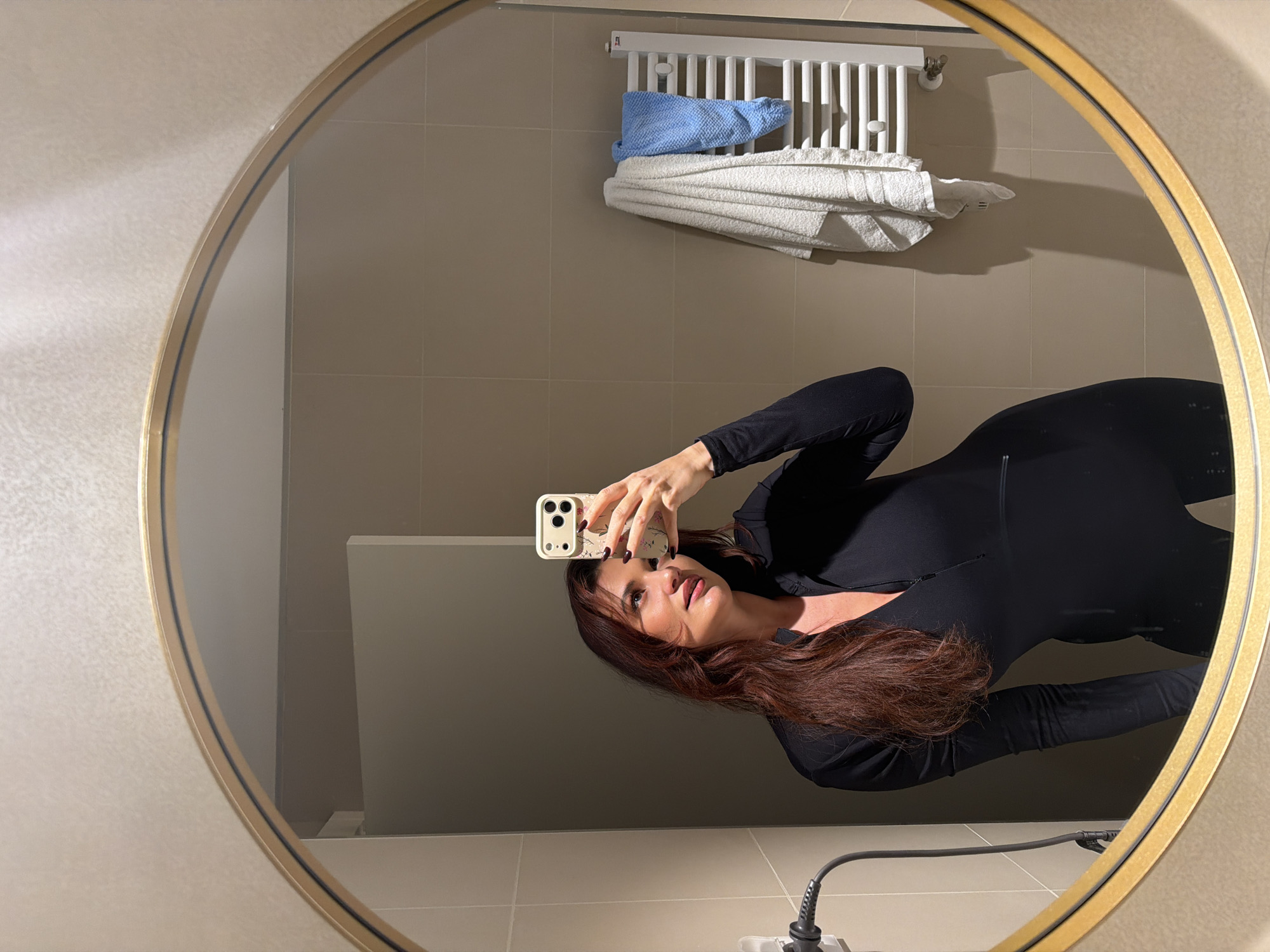 A single adult female is reflected in a large, round mirror with a gold-colored frame, capturing a selfie in an indoor bathroom environment. She has long, reddish-brown hair and is wearing a form-fitting black long-sleeved top or jumpsuit with a zippered V-neck. Her right hand, featuring dark nail polish, holds a light-colored smartphone with a floral-patterned case, positioning it for the photograph. Her gaze is directed slightly upwards and to her left, with a subtle expression. The reflected background reveals walls tiled with light beige rectangular tiles, with a lighter, textured surface above. To the right, a white horizontal bar towel radiator displays one white and one blue towel. To the left, a white electrical outlet with a plugged-in black cable is visible. A light-colored partition or door is partially reflected in the upper left. No specific visual information within the image identifies a precise location or address within the city of Belgrade, Serbia.