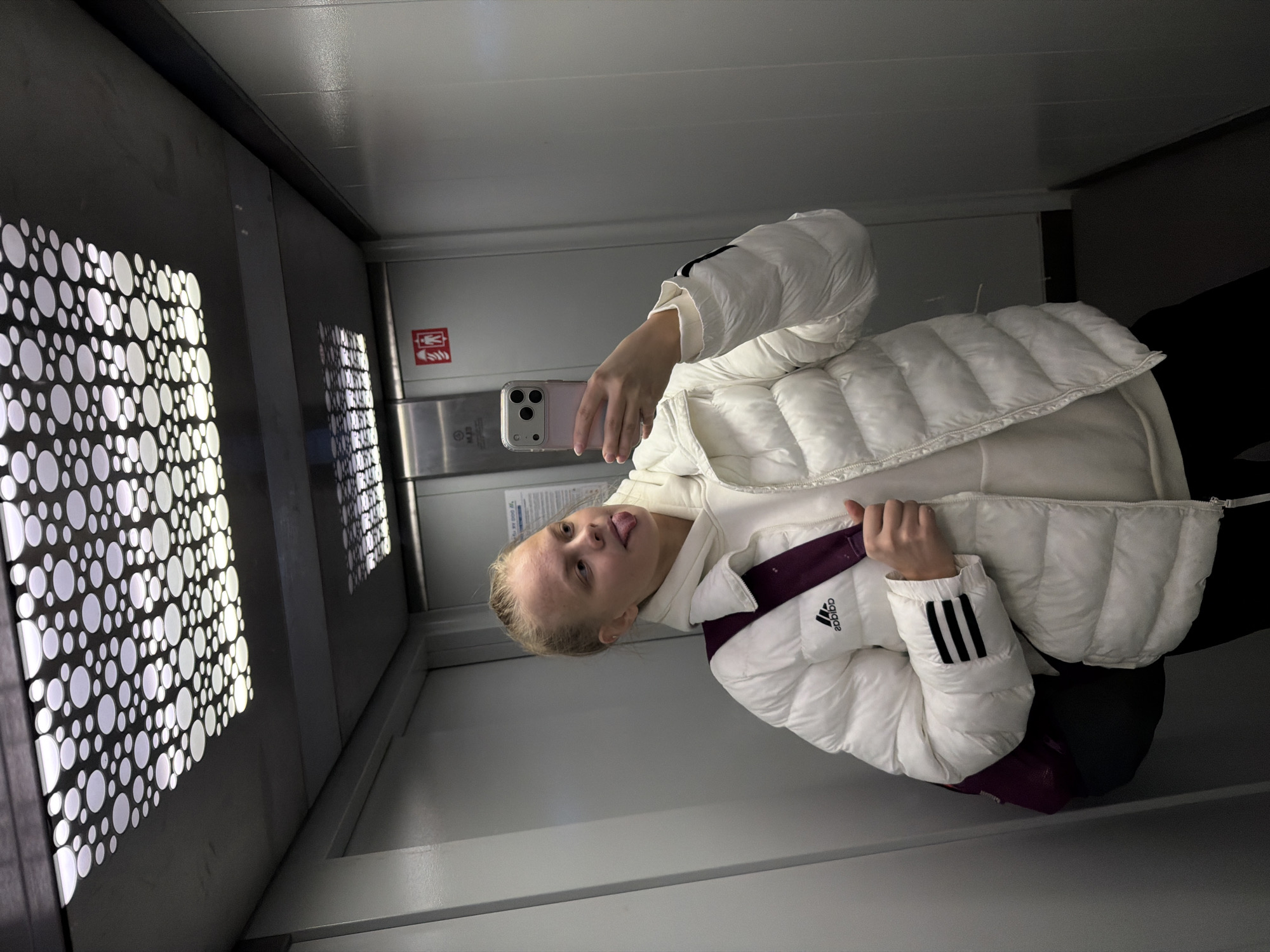 The image captures a young person, appearing to be a female with blonde hair styled back, taking a mirror selfie inside what appears to be an elevator or a modern public transport cabin in Izhevsk, Russia. She is dressed in a white puffy jacket featuring the Adidas logo on the left chest and three black stripes on the left sleeve, worn over a white hooded sweatshirt. Her expression is playful, as she is seen sticking out her tongue while holding a light-colored smartphone with multiple cameras to take the picture.
The setting is characterized by contemporary interior design. The walls of the space feature striking panels with illuminated white circular patterns against a dark background, creating a decorative and modern ambiance. Adjacent to these panels, there is a metallic surface and a white wall, suggesting a blend of materials. A sign is visible on one of the walls, displaying a red symbol commonly associated with fire safety (a flame and a figure), beneath which there is some indistinct Cyrillic text. The lighting within the space is artificial, casting a bright glow on the subject and the immediate surroundings. The overall mood is casual and lighthearted, captured through the selfie activity and the subject's playful pose.