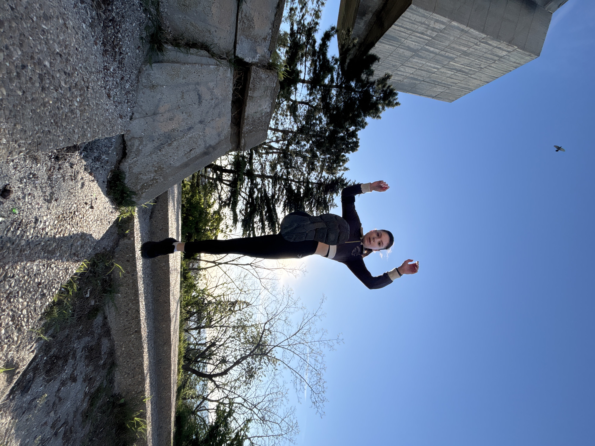 A young adult female is photographed at the Monument of the Soviet-Bulgarian Friendship in Varna, Bulgaria. She wears dark leggings, a long-sleeved top, and dark footwear, along with what appears to be a weighted vest. She is positioned horizontally against the clear blue sky, with the monument's architectural elements visible in the background. Trees, shrubs, and vegetation surround the monument's concrete structure. The sun's glare is present, particularly behind the trees. A bird is visible in the upper right portion of the sky.
