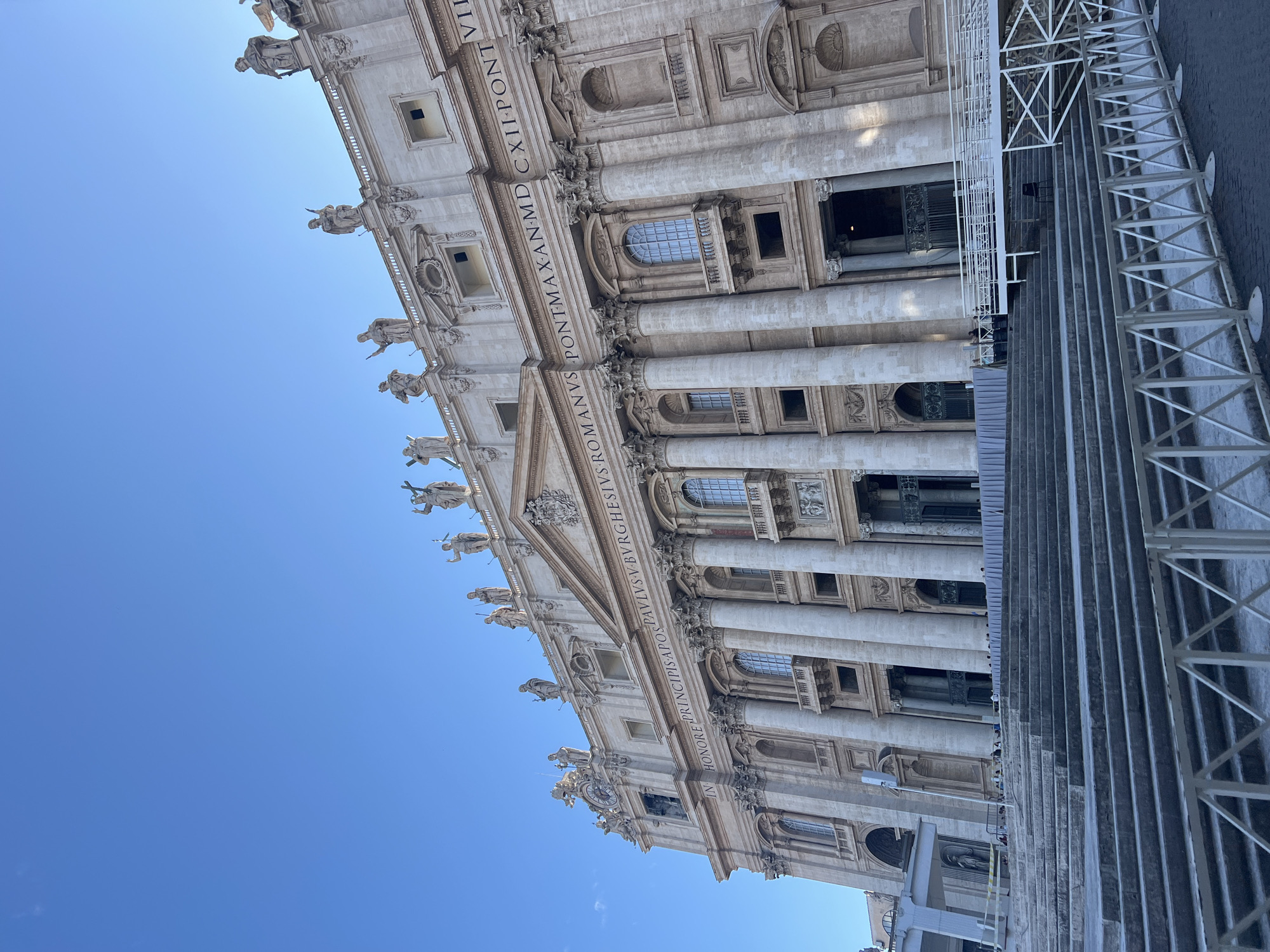 The image displays a low-angle view of the facade of St. Peter's Basilica in Città del Vaticano, Italy, under a clear blue sky. The grand Baroque facade, constructed primarily of travertine, features multiple levels.
The upper section of the facade is adorned with a balustrade supporting numerous large, detailed marble statues of saints, including figures holding a cross and other attributes, silhouetted against the sky. Below the statues, a prominent entablature features Latin inscriptions. On the visible left, the inscription reads "IN HONORE PRINCIPIS APOS," and on the visible right, "PAVLVS V BVRGHESIVS ROMANVS PONTMAX AN MDCXII PONT" is partially visible.
The mid-section of the facade is characterized by massive Corinthian columns, with recessed arched windows and balconies positioned between them. On the far left, a clock face is visible within the architectural details. Multiple ornate, dark-colored entrance doors are visible at the base of the facade.
In the foreground, a wide, multi-tiered set of stone steps leads up to the basilica's entrances. These steps show signs of wear. White metal barricades with an X-pattern design span the immediate foreground, separating the viewer from the steps. A white temporary ramp structure, supported by scaffolding, extends from the right side of the steps towards one of the entrances. A single black chair is situated on the steps beneath this ramp. On the far left, another white temporary structure, possibly a stage or covered area, is present with a small statue underneath it. A small number of individuals are visible on the steps and near the entrances, appearing to be queuing or gathered. The paved ground beneath the barricades is dark. The scene is brightly illuminated by natural light, casting distinct shadows.