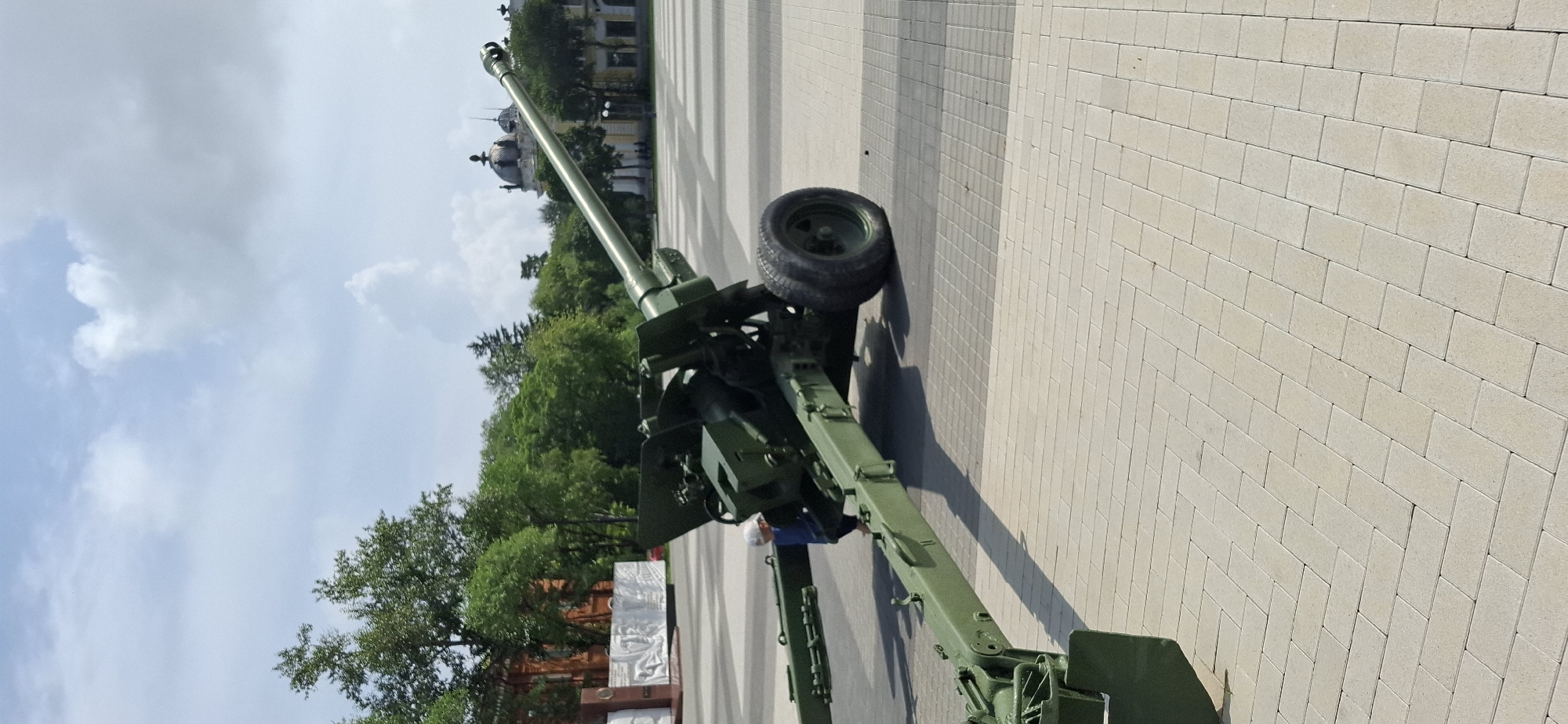 A military cannon, painted olive green, is positioned on a paved plaza. The cannon is angled upwards and to the right. A person wearing a white hat and a blue shirt is visible near the center of the cannon's base. In the background, trees line the edge of the plaza, and several buildings are visible, including one with a domed roof. The sky above is partially cloudy. This scene is located in Blagoveshchensk, Russia, likely at a memorial or historical site, given the presence of the cannon and the surrounding architecture. The paved area is made of light-colored rectangular bricks laid in a patterned fashion.