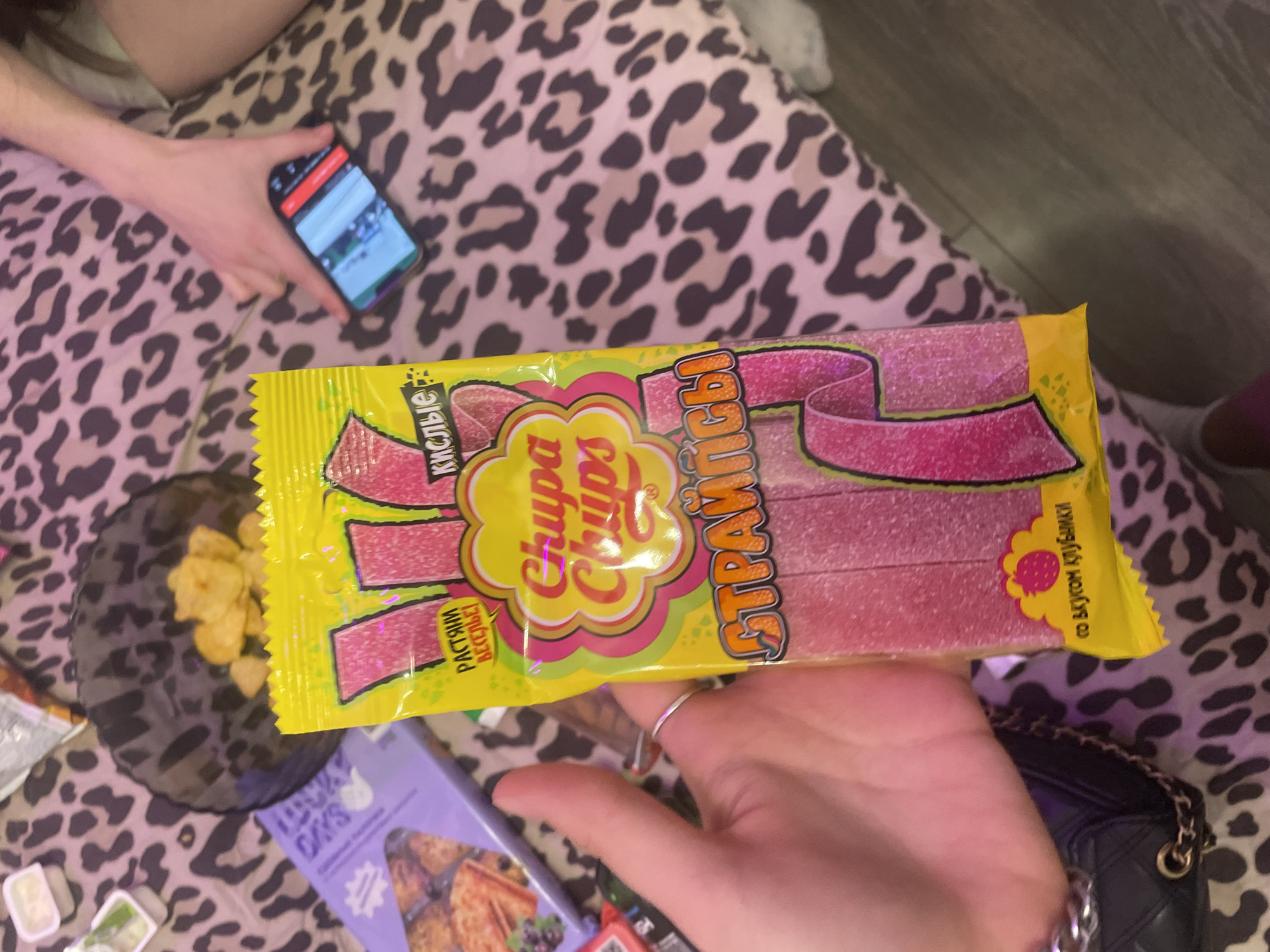 In the city of Zmeinyi, Russia, a person is holding a yellow package of "Chupa Chups" sour straws. The package is pink and features the Chupa Chups logo. The straws themselves are also pink and appear to be sprinkled with sugar. In the background, a hand is visible holding a smartphone, and a leopard-print blanket covers the surface. There are also some chips in a black bowl and a purple box in the background.