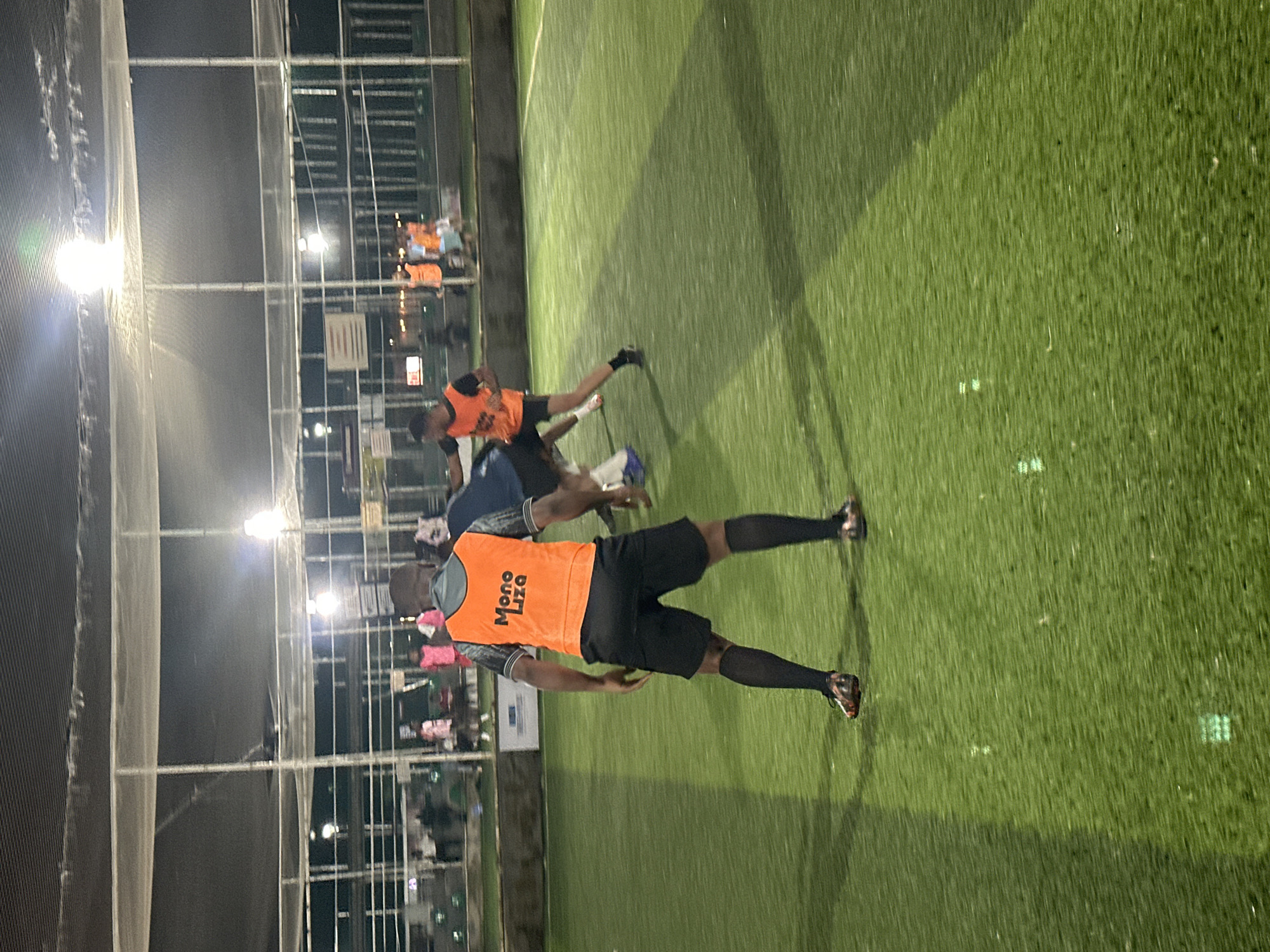 The image depicts an outdoor artificial turf soccer field at night, illuminated by overhead lights. The scene is set in Abuja, Nigeria. In the foreground, a person wearing an orange vest with "Mono Liza" written on it is facing away from the camera, standing on the field. This individual is wearing black shorts and black socks. To the right of this person, another individual in a blue shirt and white shorts is engaged in a soccer game, with their left leg extended. In the background, several other individuals are visible on the field, some wearing orange vests and others in civilian clothing. The field is bordered by a fence and netting, with buildings and lights visible beyond the fence. The overall atmosphere suggests a casual soccer match or training session taking place after dark.