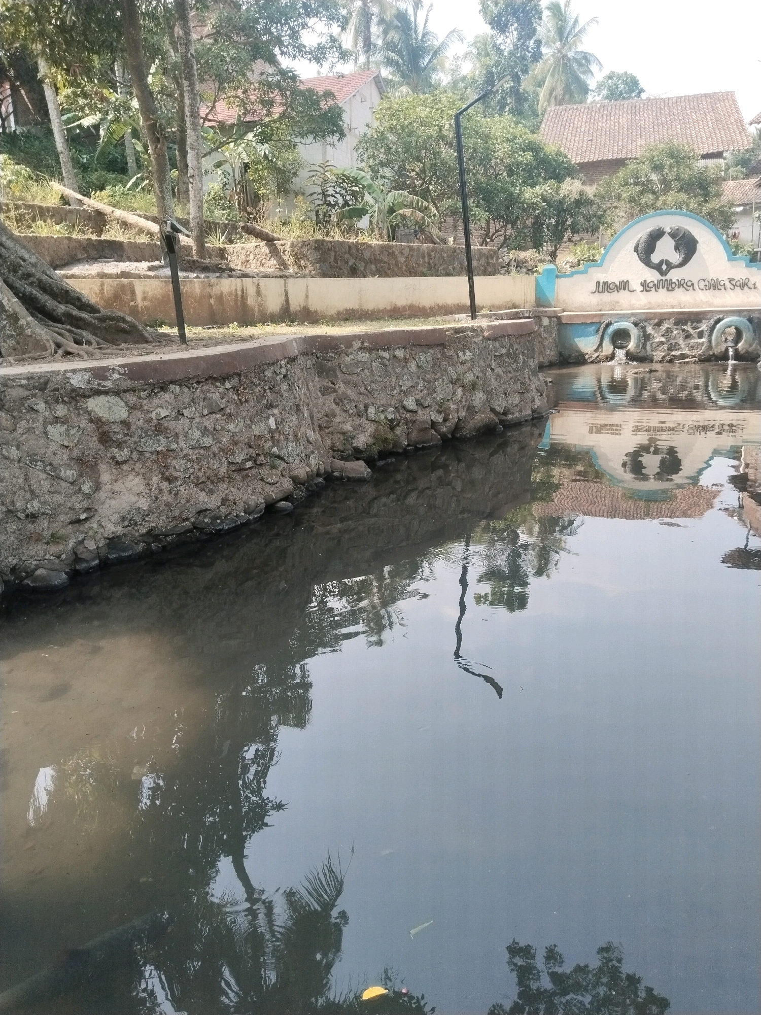 The image shows a stone-walled canal in Cenggini, Indonesia, with a body of water reflecting trees and buildings. A decorative archway with a symbol and text is visible on the far bank, above the water. Two spouts are visible, feeding into the canal. The canal wall on the left is constructed from rough, irregular stones. Trees with visible roots grow on the left bank, and vegetation is present in the background, along with houses with red tile roofs. The water's surface is mostly calm, with some ripples and reflections. A thin, dark object, possibly a branch or debris, is floating in the water in the center of the frame.