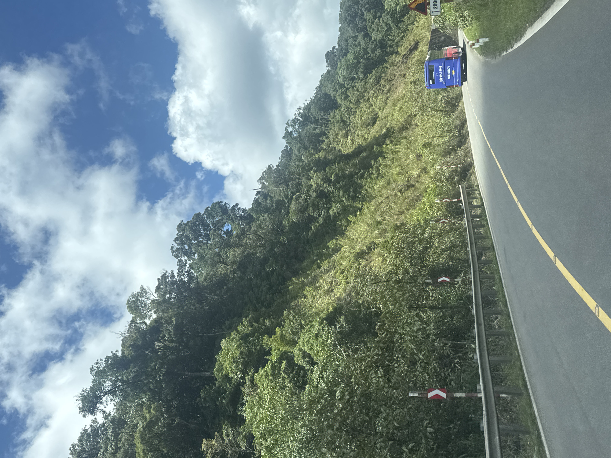 A bright, sunny day with scattered clouds over a winding, paved road in Nam Khánh Vĩnh, Vietnam. The road curves to the right, bordered by a metal guardrail on the left and a steep, densely vegetated hillside on the right. The hillside is covered in lush green trees and foliage, suggesting a mountainous or hilly terrain. Along the roadside, there are several warning posts with red and white directional arrows, indicating a curve in the road. In the distance, a blue tour bus with white lettering on its side and a red vehicle are visible driving away from the camera. A yellow line marks the center of the road. A yellow triangular road sign with a black border is visible on the right side of the road, along with a white sign indicating "200m." The overall scene depicts a scenic drive through a natural landscape.