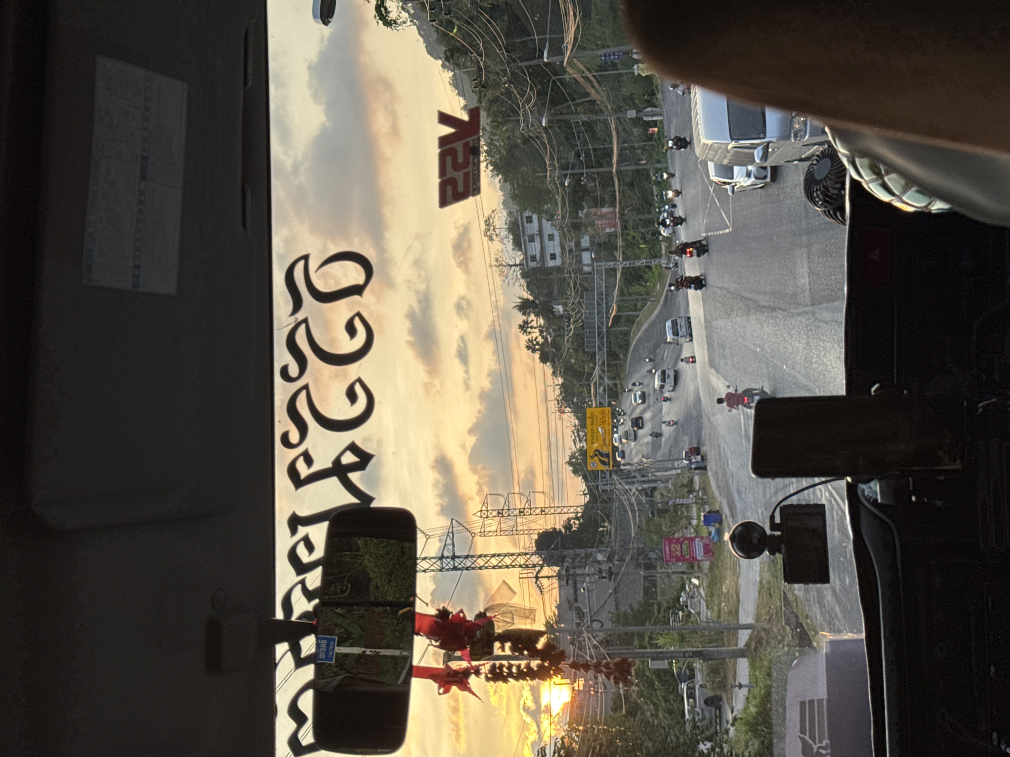 The image depicts a view from inside a vehicle, likely a bus or truck, moving along a road in Patong, Thailand, during what appears to be late afternoon or early evening. The outside view is seen through the windshield, with a portion of the vehicle's interior visible at the edges of the frame. The road has moderate traffic, including cars, motorcycles, and possibly other vehicles. Utility poles and wires line the roadside, along with trees and some buildings. The sky shows a sunset or late afternoon glow with scattered clouds. Reflections on the windshield obscure some of the details. A sign with "22V" is visible on the building.