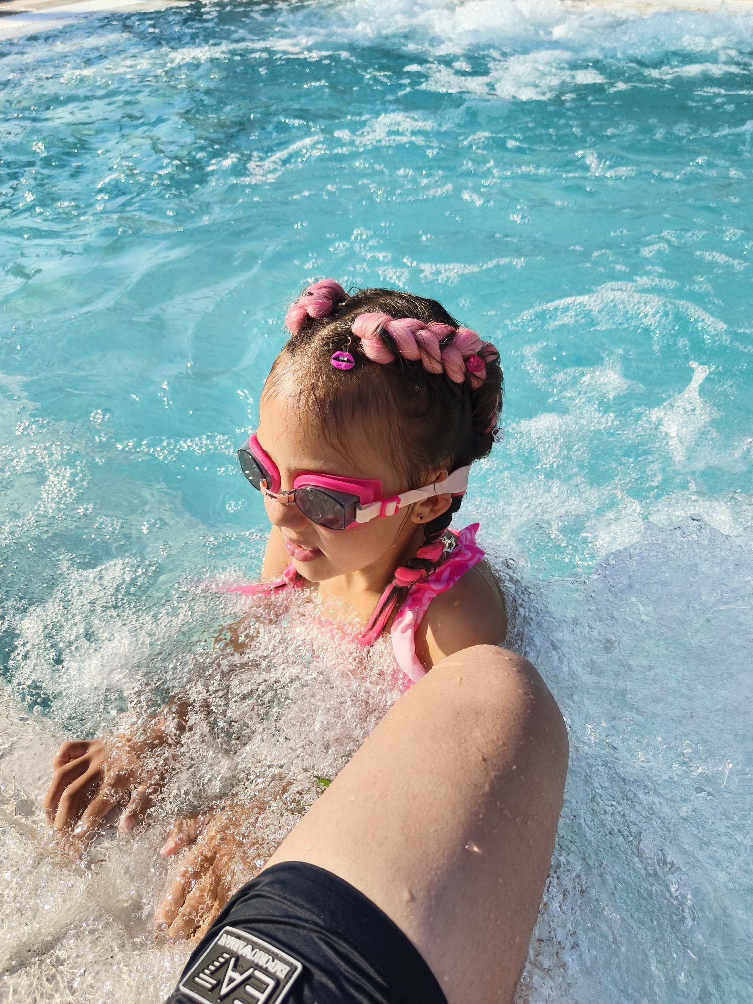 This media file features a young girl with pink braided hair, adorned with pink lip-shaped charms, wearing pink swimming goggles and a pink swimsuit. She is partially submerged in clear blue water, which appears to be a swimming pool or a hot tub, indicated by the bubbling water and foam around her. The water is a vibrant turquoise color, with sunlight creating shimmering patterns on its surface. The girl is looking to her right with a slight smile, suggesting enjoyment or anticipation of a splash. Her hands are visible in the water, creating ripples and splashes. In the foreground, a person's leg, wearing black swim trunks with the "EA7 EMPORIO ARMANI" logo, is visible. The lighting suggests it is a sunny day, likely during daylight hours. The overall scene conveys a sense of fun and relaxation in a water-based activity, possibly a vacation in Kiten, Bulgaria.