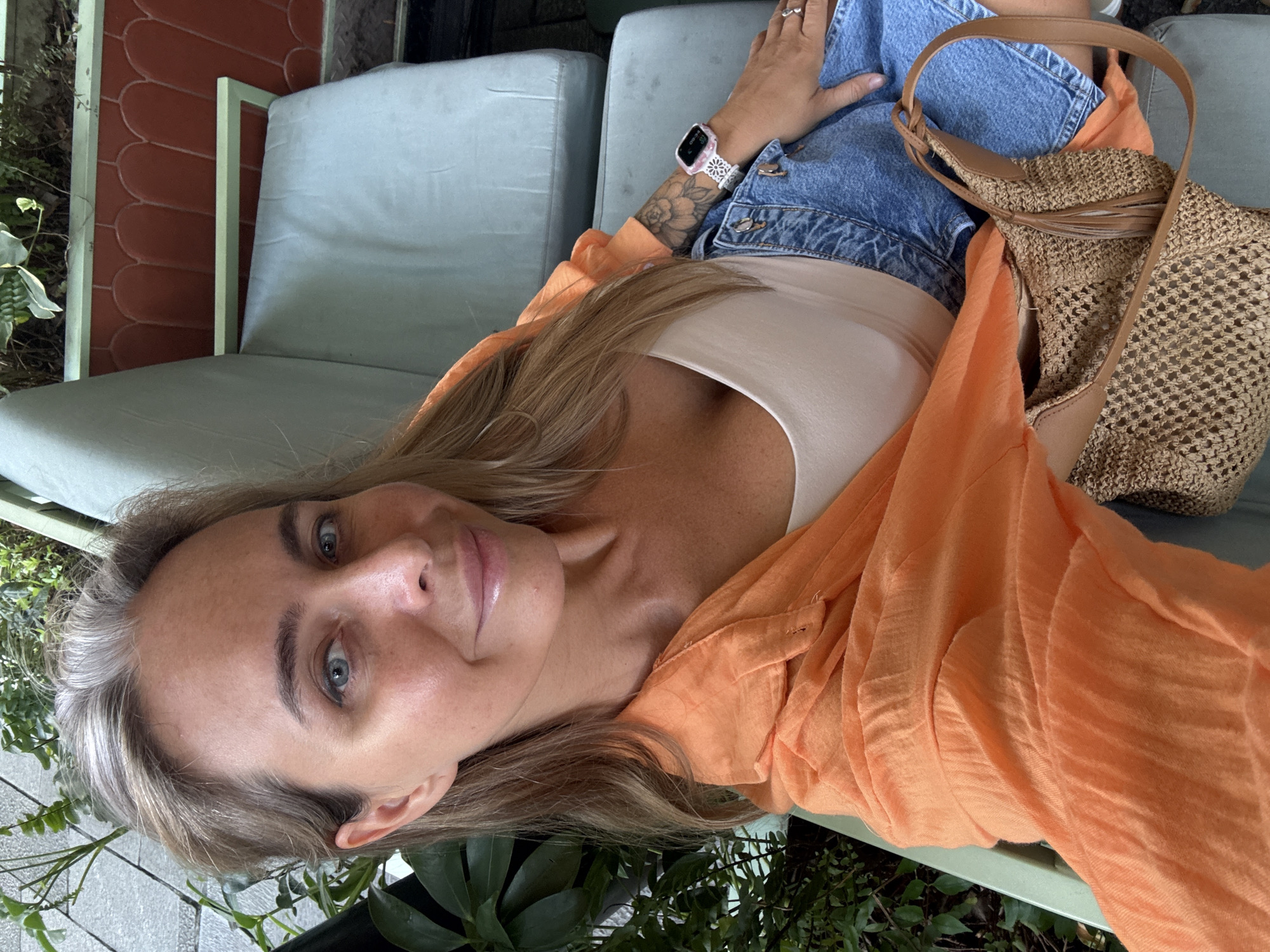 A woman with blonde hair, blue eyes, and grey streaks is sitting on a light blue chair outdoors. She is wearing an orange open shirt, a beige tank top, and denim shorts. She has a tattoo on her left arm and is wearing a watch on her left wrist. A woven handbag with leather straps is on her lap. She is looking at the camera and smiling. The background shows greenery, a reddish-orange tiled wall with arched patterns, and more light blue chairs. The setting appears to be a cafe or restaurant patio.