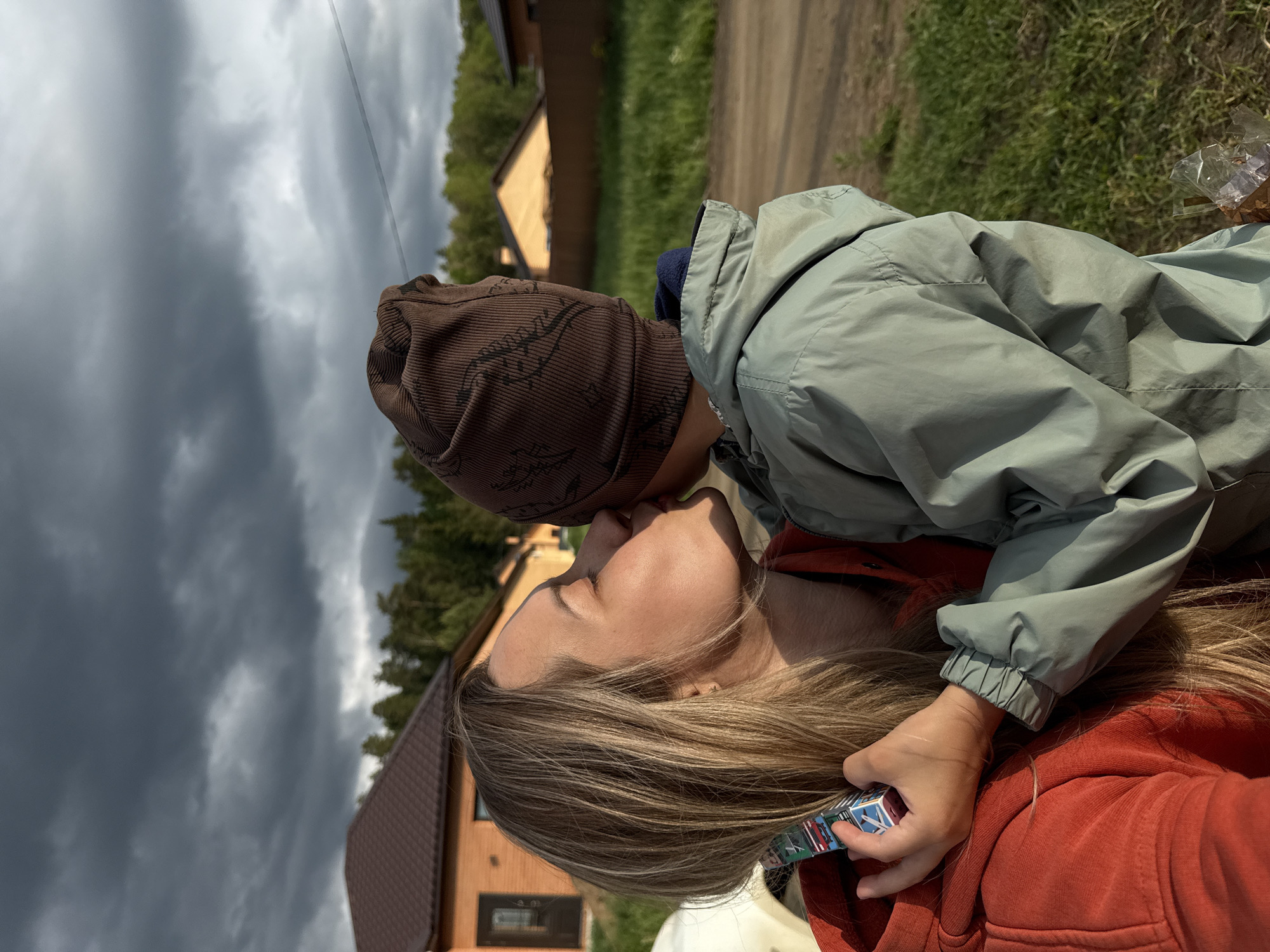 A woman and a child are outdoors. The woman is wearing a red hoodie, and the child is wearing a brown hat and a green jacket. The child is kissing the woman on the nose. The woman is holding a small rectangular object in her hand. In the background are houses and trees. The sky is cloudy. The location is Chaikovsky, Russia.