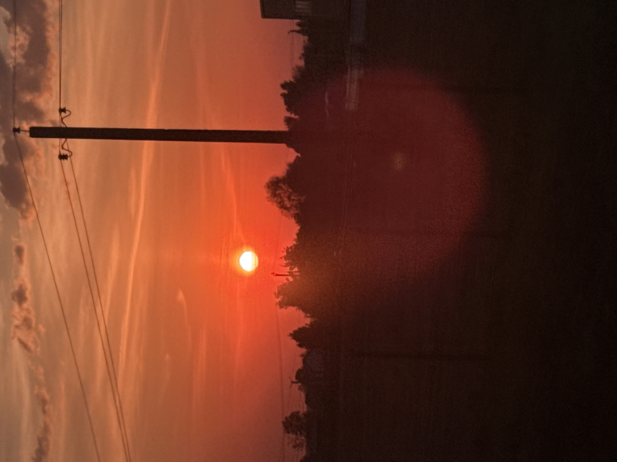 The image captures a sunset over Nikol'skoe, Russia. An orange sun is positioned low in the sky, partially obscured by trees. A utility pole with power lines is visible in the foreground. The sky displays streaks of orange and pink, suggesting a hazy atmosphere. A dark, undefined mass is present on the right side of the image.