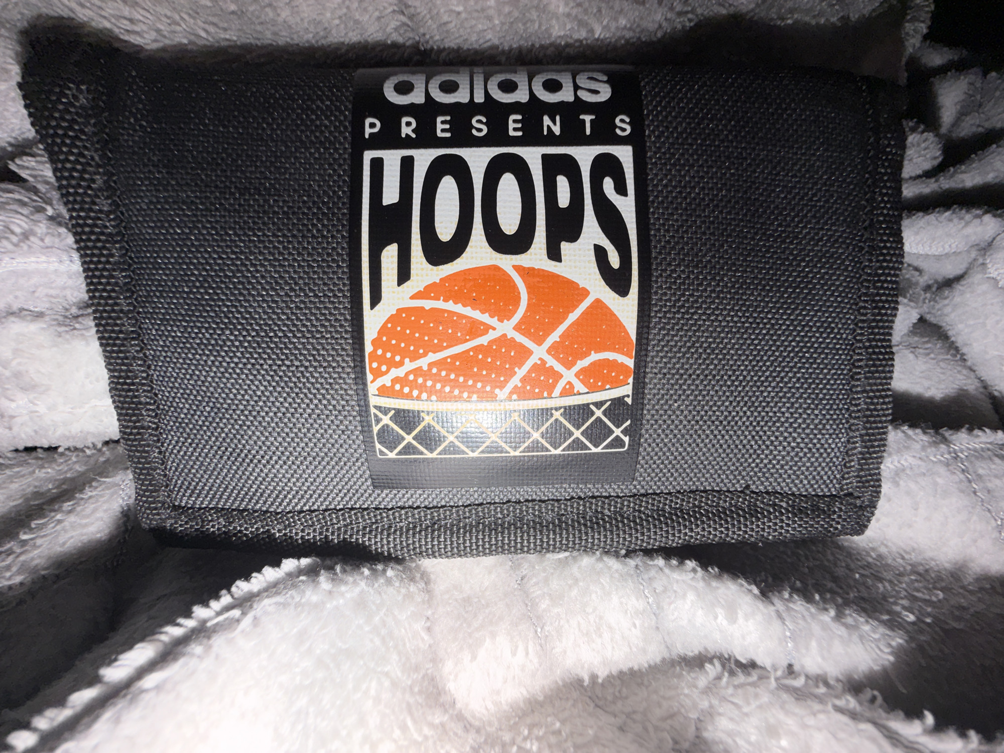 The image shows a close-up, overhead view of a dark grey fabric object, possibly a wallet or pouch, with an "Adidas Presents Hoops" logo. The logo features the Adidas name in white, followed by "PRESENTS" in smaller white text, and "HOOPS" in large black letters. Below "HOOPS" is a graphic of an orange basketball with white dots and black lines, and a white net pattern. The object is placed on a soft, white, fluffy surface. The image does not contain any information about the city of Dorsten, Germany.