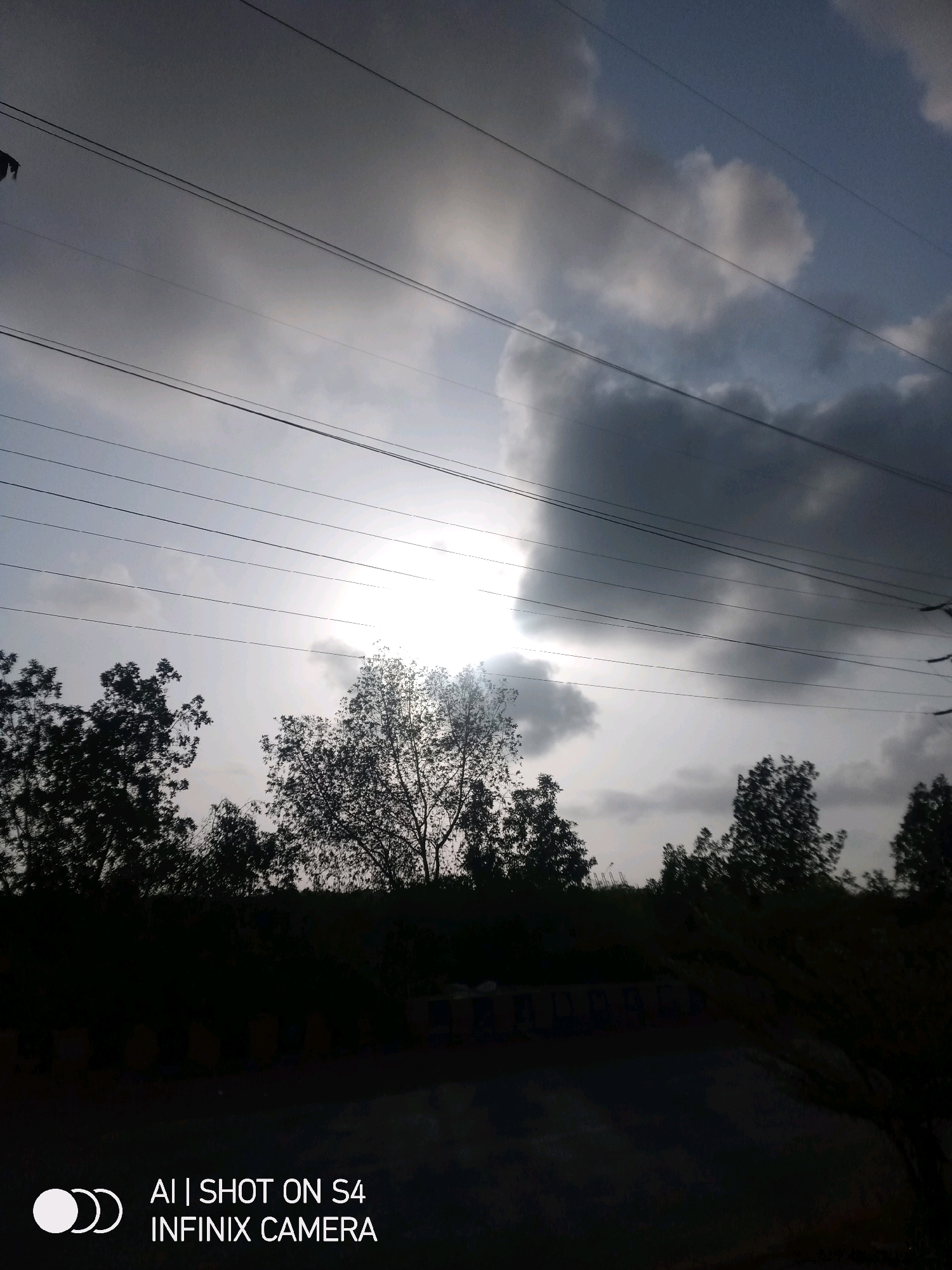 The image depicts an outdoor scene with a cloudy sky and several parallel power lines running horizontally across the frame. Silhouetted trees line the bottom of the image, with their dark forms contrasting against the lighter sky. The sun is visible behind a dark cloud, creating a bright, hazy effect. The lower portion of the image shows what appears to be a road or pathway and some indistinct structures or markings. Text overlays at the bottom right corner read "AI | SHOT ON S4 INFINIX CAMERA". While the image is set outdoors, the specific location within Karachi Division, Pakistan, cannot be determined from the visual information provided.
