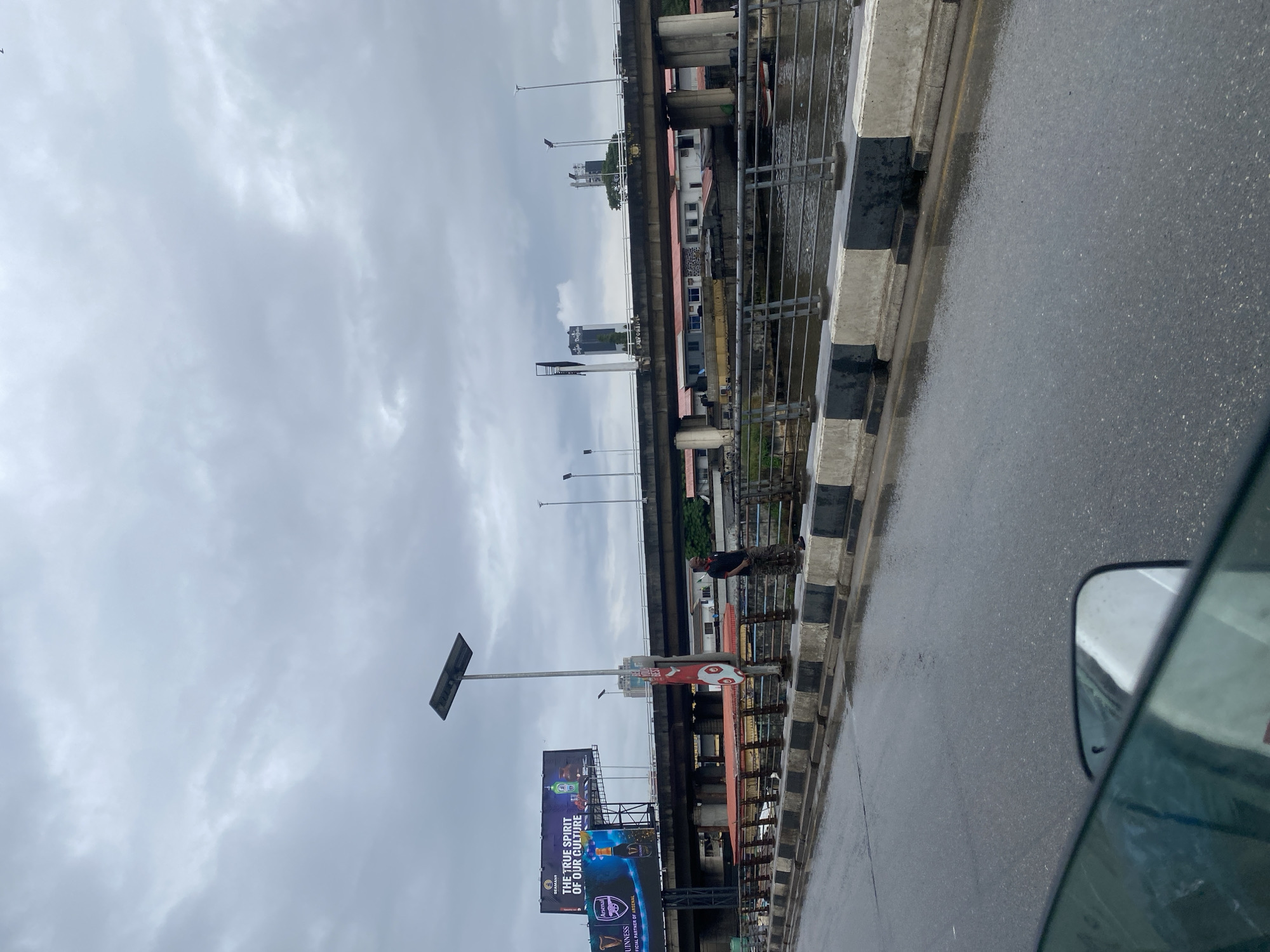 The image depicts a wet road with a man standing on the sidewalk next to a concrete barrier. A large billboard displaying an advertisement for Guinness is visible in the background, along with an elevated highway and a body of water. The sky is overcast with clouds, suggesting it may have recently rained or is about to rain. The scene appears to be in a city, possibly Victoria Island, Nigeria, given the presence of billboards and urban infrastructure. The man is dressed casually in a dark t-shirt and camouflage pants. The overall atmosphere is one of a typical urban environment under cloudy skies.