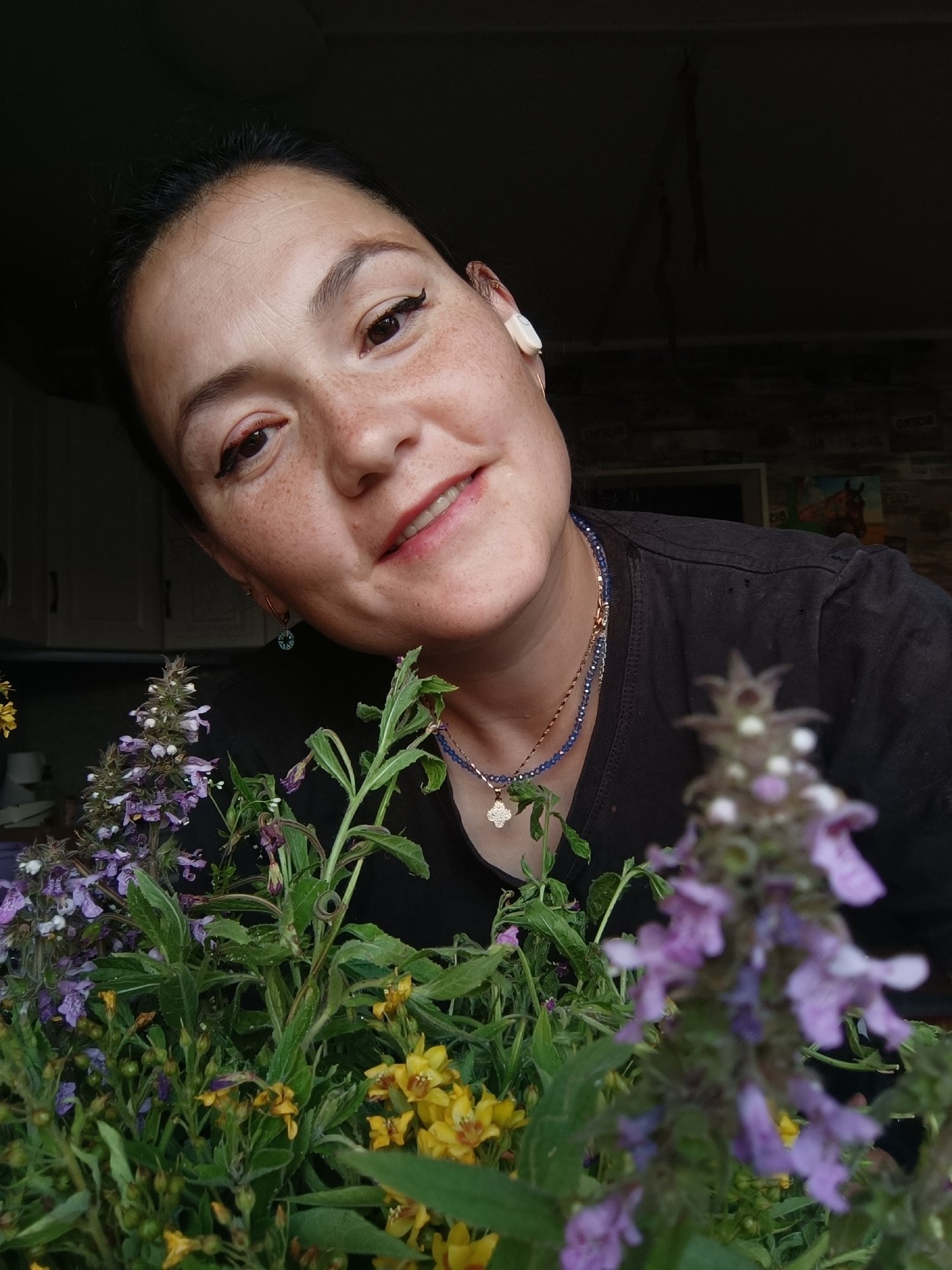 A close-up, high-angle shot shows a woman with light brown hair, dark brown eyes, and freckles smiling. She is wearing a black t-shirt, a white wireless earbud in her right ear, and a necklace with blue beads and a four-petal clover pendant. In the foreground, out of focus, are various wildflowers, including purple and yellow blooms. The background shows a partially visible kitchen area with cabinets and a framed picture on the wall. The scene appears to be indoors.