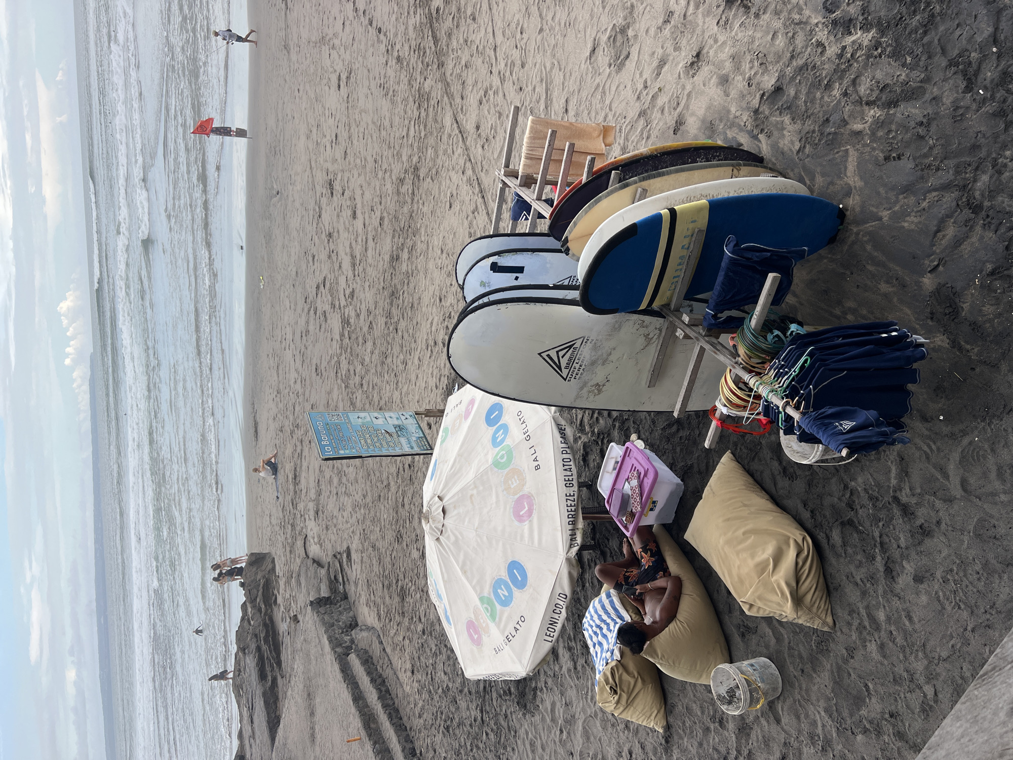 This image captures a vibrant beach scene in North Kuta, Indonesia, likely during the daytime under a partly cloudy sky. The dark, possibly volcanic sand of the beach fills the majority of the frame, with the ocean visible on the left.
In the foreground, a large white beach umbrella, advertising "BALI GELATO," "LEONI.CO.ID," and "BALI BREEZE, GELATO PLEASE!", provides shade. Beneath it, a person relaxes on a beige beanbag chair, seemingly engrossed in a phone. Another beanbag chair and a plastic cooler with a pink lid are also visible nearby, along with a bucket on the sand.
To the right of the umbrella, a stack of diverse surfboards, including white, blue, yellow, and black ones, are propped up on wooden racks. A sign on one of the white surfboards reads "SURF LESSONS BARUNA PERERE". Adjacent to the surfboards, a wooden stand holds neatly folded blue towels and what appear to be ropes or leashes, suggesting a rental or surf school setup. Another blue sign, visible further back, also advertises "La Baruna SURF LESSONS" and "BEGINNER CLASS".
In the mid-ground and background, the ocean stretches out with gentle waves. Several people are scattered along the shoreline; some wade in the water, while others stand on rock formations. A person is seen kite surfing in the distance, and another individual is visible near the water's edge with a red flag or banner. The atmosphere appears leisurely and inviting, typical of a popular beach destination.